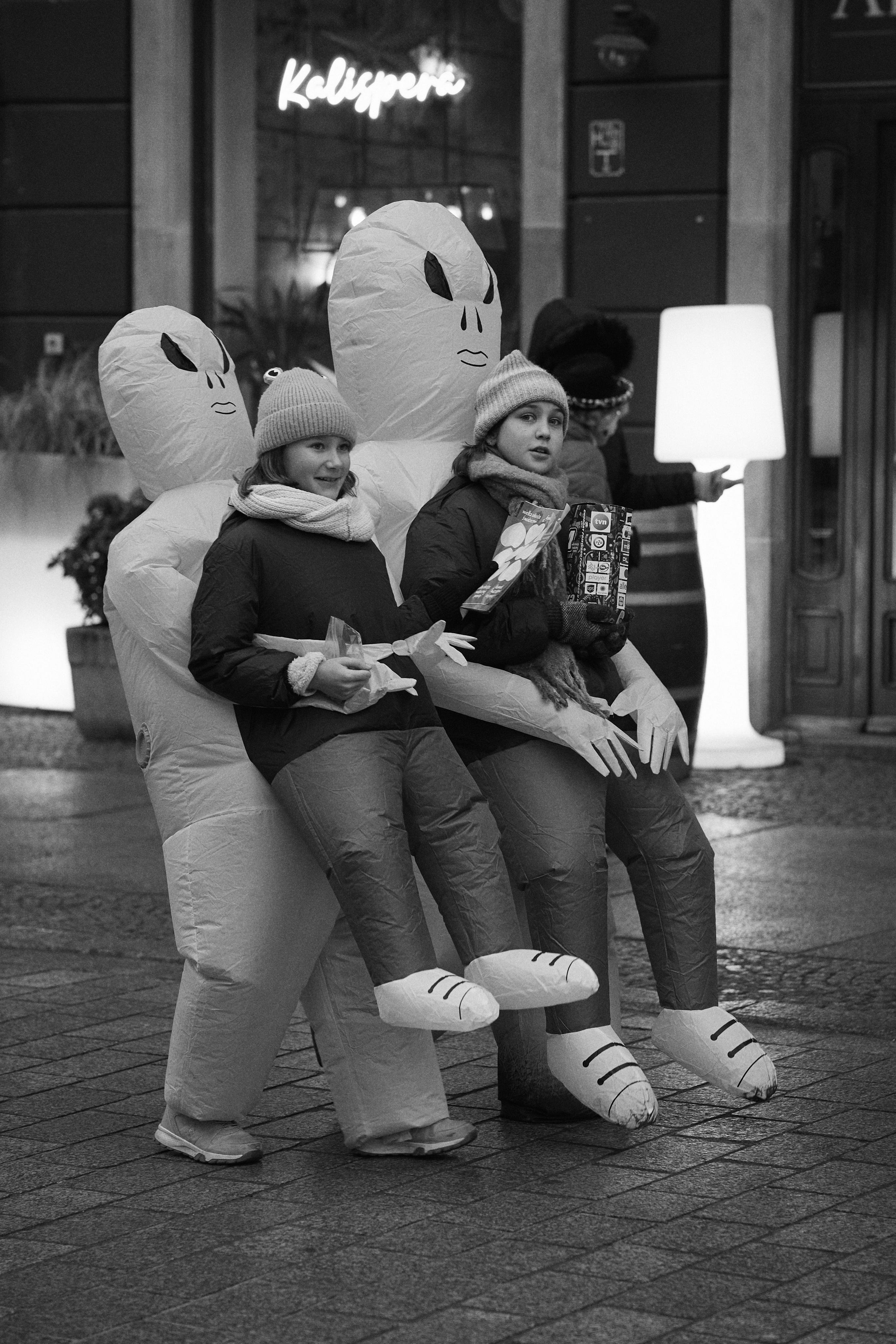 Two children in winter coats and knit hats pose on a sidewalk wearing inflatable alien costumes that make it look like the aliens are carrying them.