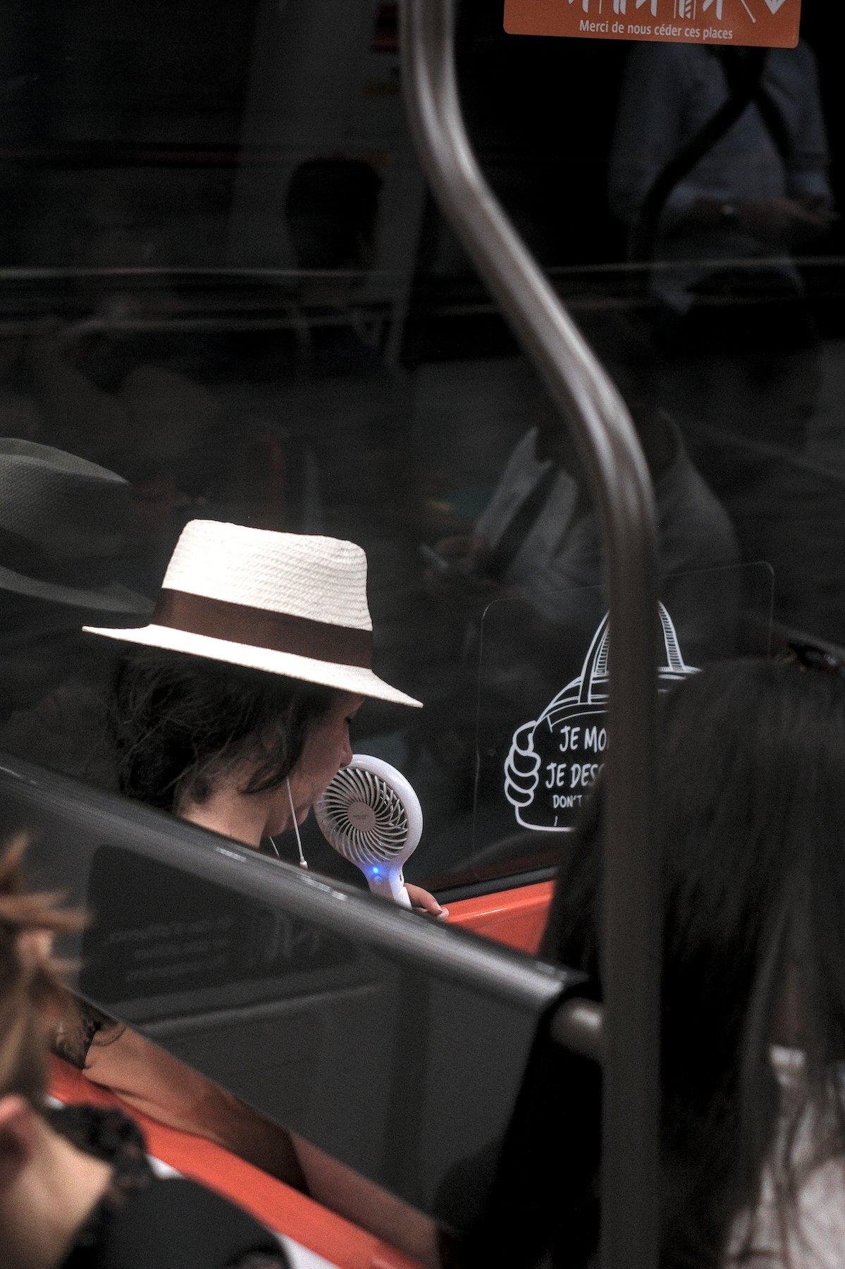Photo verticale. Dans le métro, assise à un carré de 4, une femme de 3/4 dos, aux cheveux noirs, avec un chapeau élégant blanc et un mini ventilateur portatif blanc