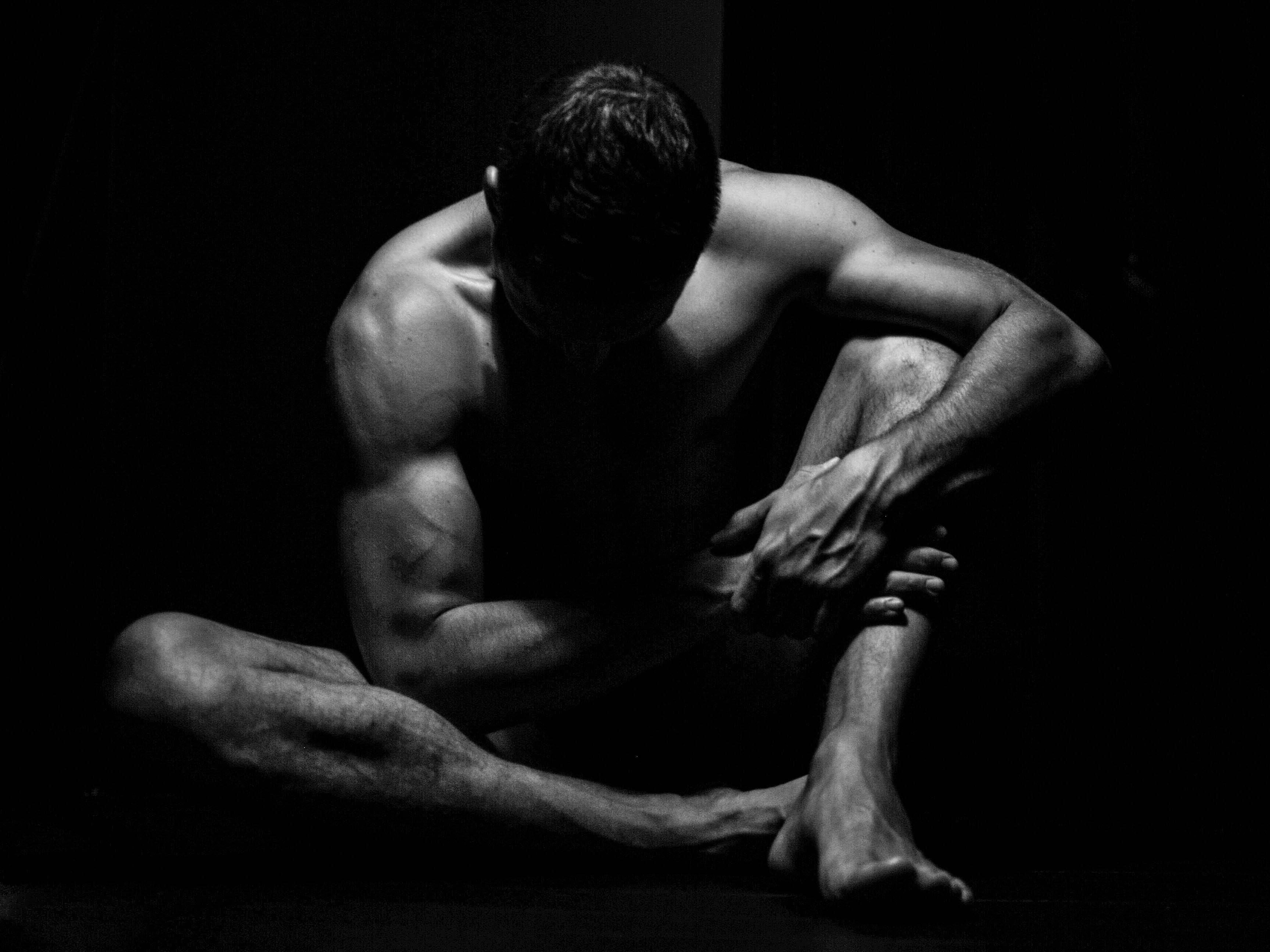 This black and white image shows a shirtless person sitting on the floor in a contemplative pose. The lighting emphasizes their muscular build, creating a dramatic and artistic effect with shadows. The person is leaning forward with their arms resting on their legs.