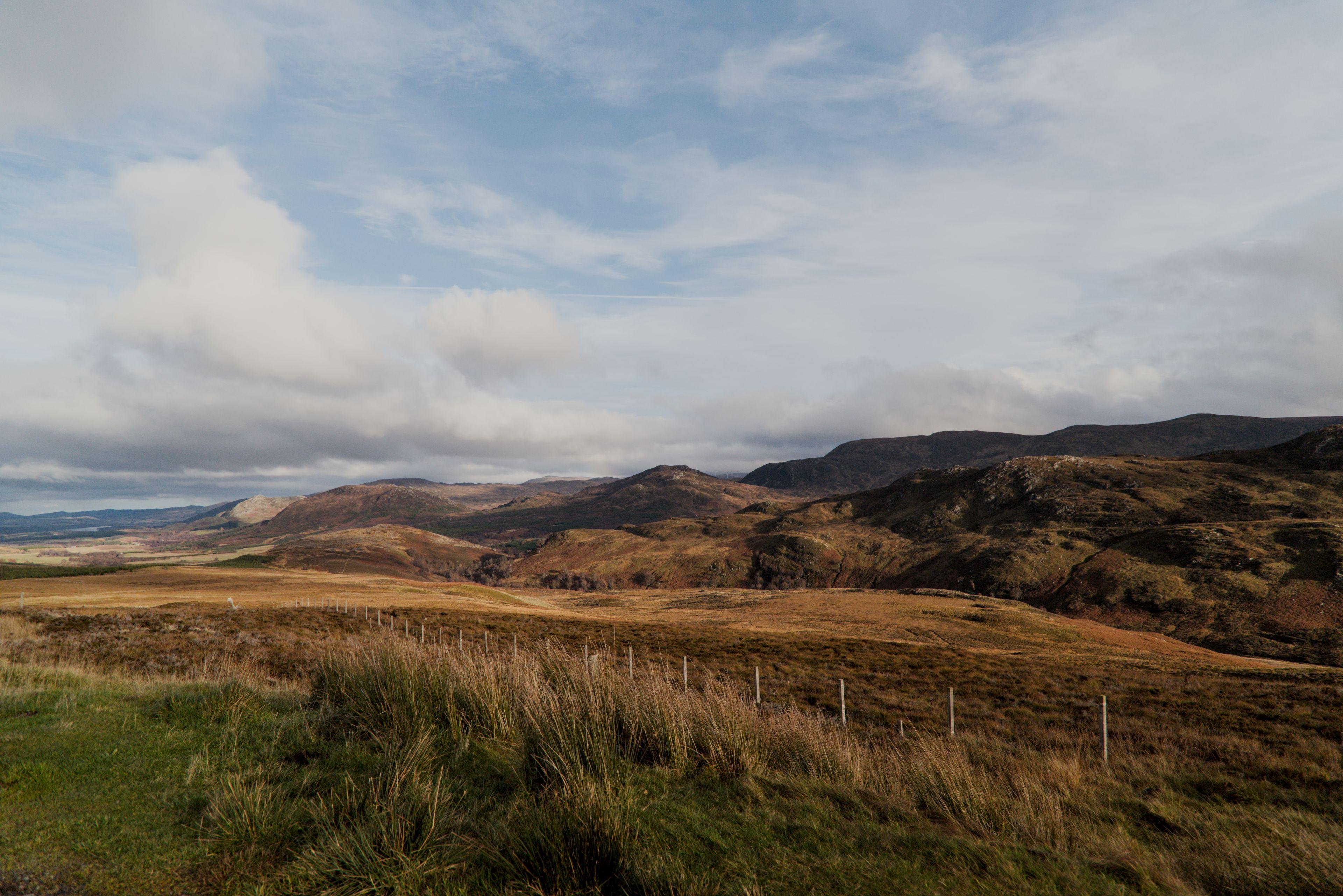 Photo format paysage montrant les Highlands écossais, un morceau d’herbe verte au premier plan, puis le reste du sol est dans les teintes marrons, avec des reliefs au fond, sous un ciel bleu mais très nuageux