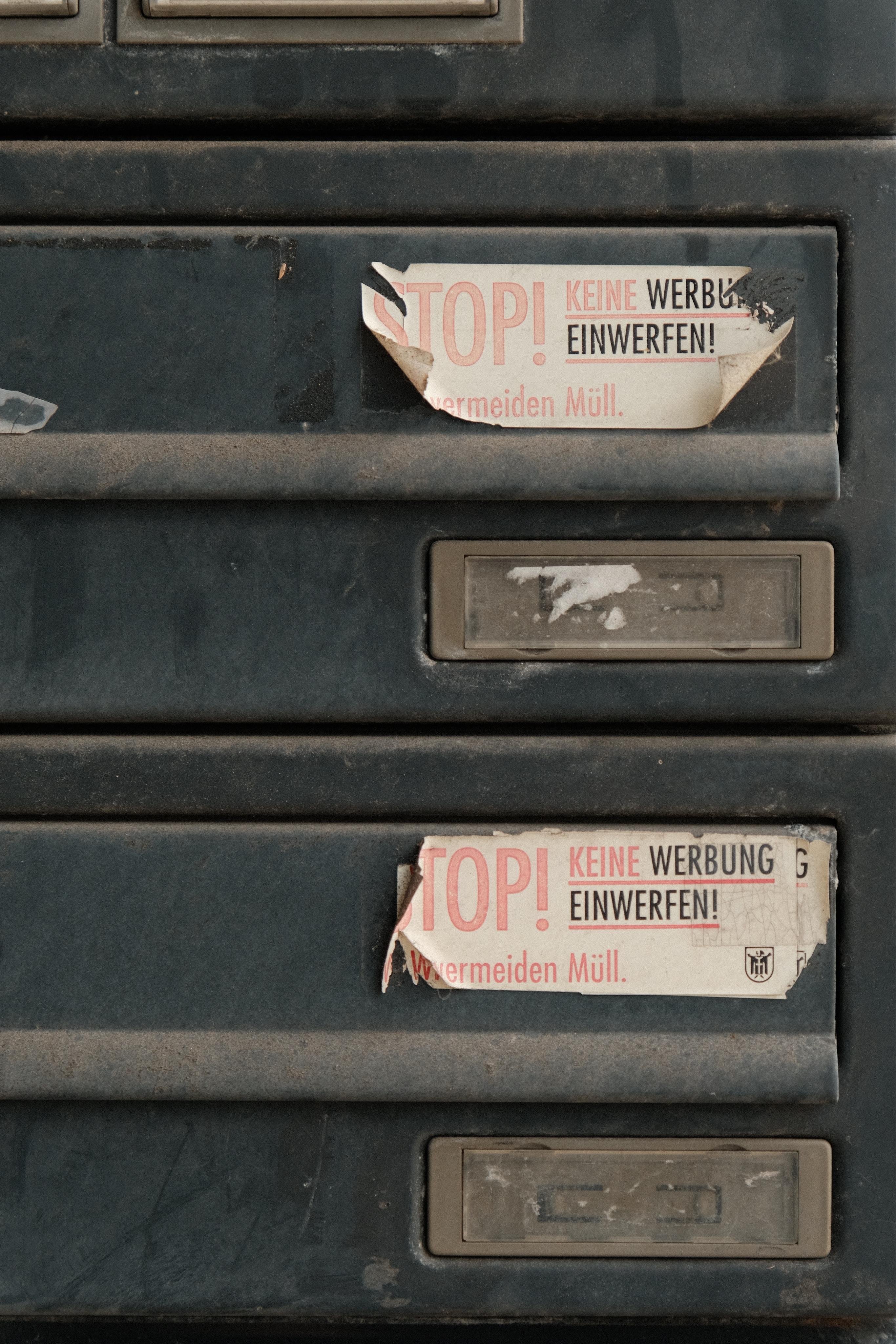Close-up of dark metal mailboxes with stickers reading “STOP! KEINE WERBUNG EINWERFEN!” and empty label slots below. The stickers are torn, the metal shows dirt and wear.