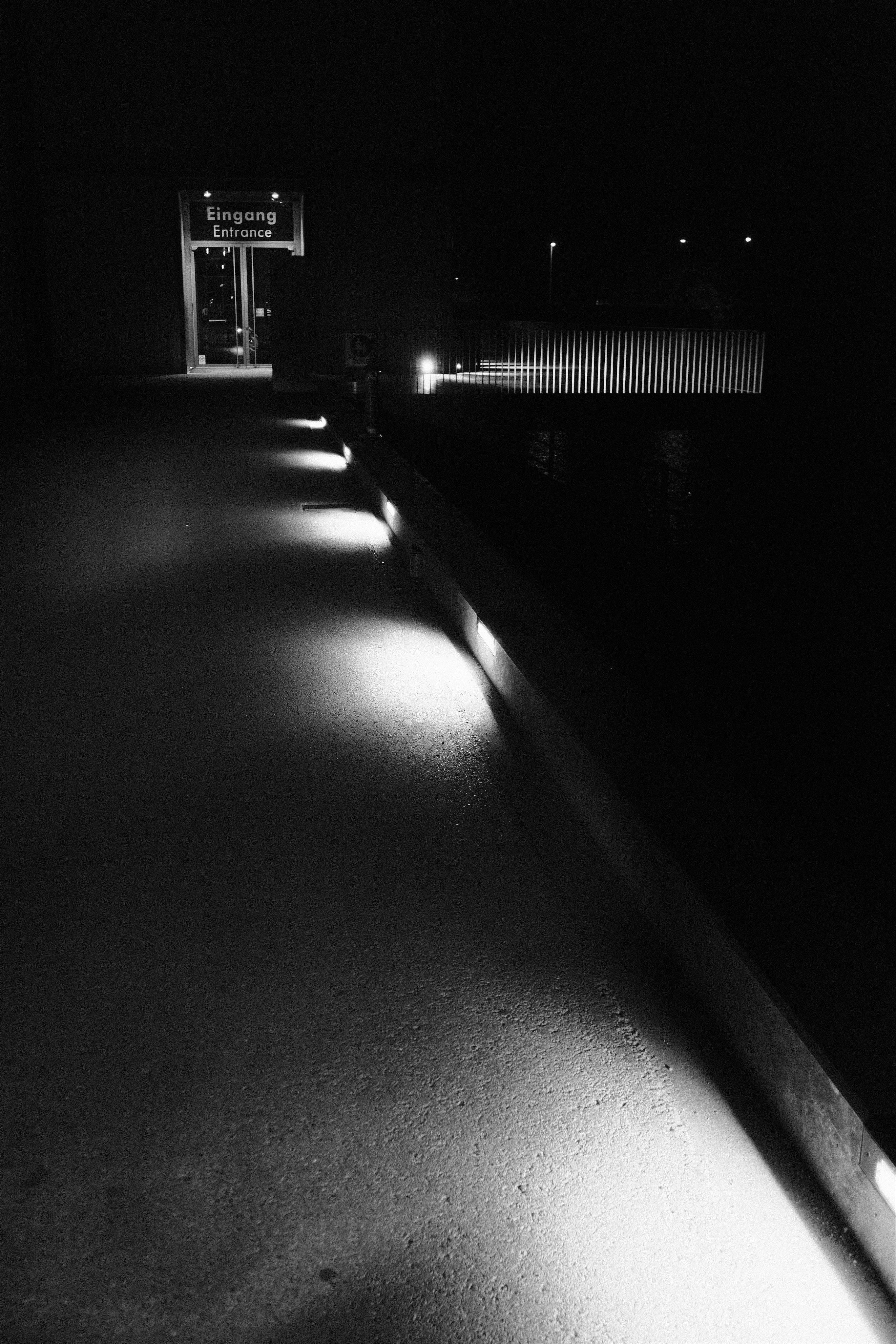 Monochrome image. A dark asphalt walkway illuminated by 5 low-lying lamps leading up to an illuminated door with a sign above saying „EINGANG“ and „ENTRANCE“. The rest of the image is pitch black, so you cannot see the surroundings and not even the building to which the entrance is leading.