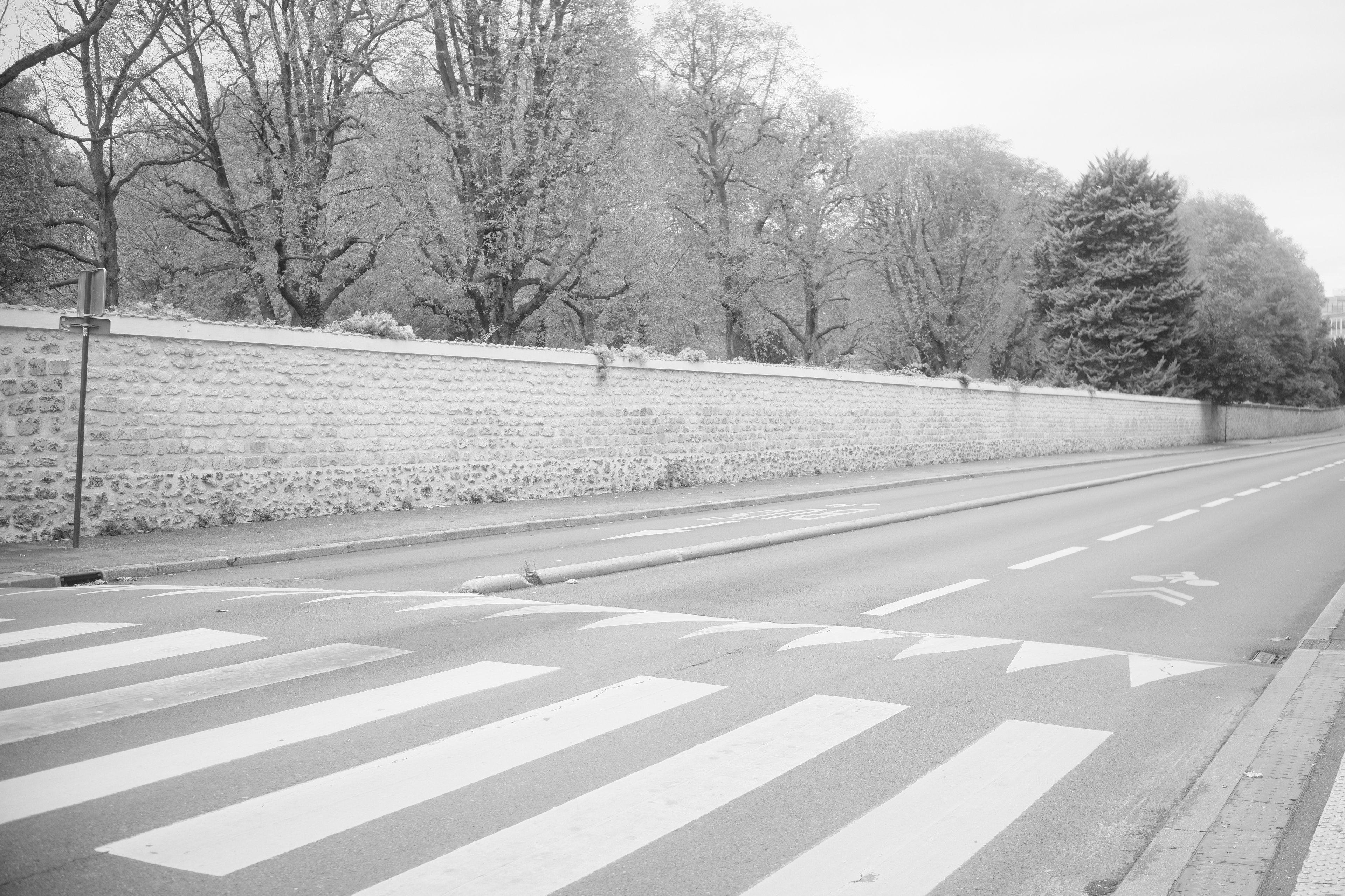 Photo horizontale en noir et blanc. Une route à deux voies, une dans chaque sens, et une voie bus protégée par une butée. Un passage piéton au premier plan, un mur de pierre tout le long de la rue et des arbres derrière le mur