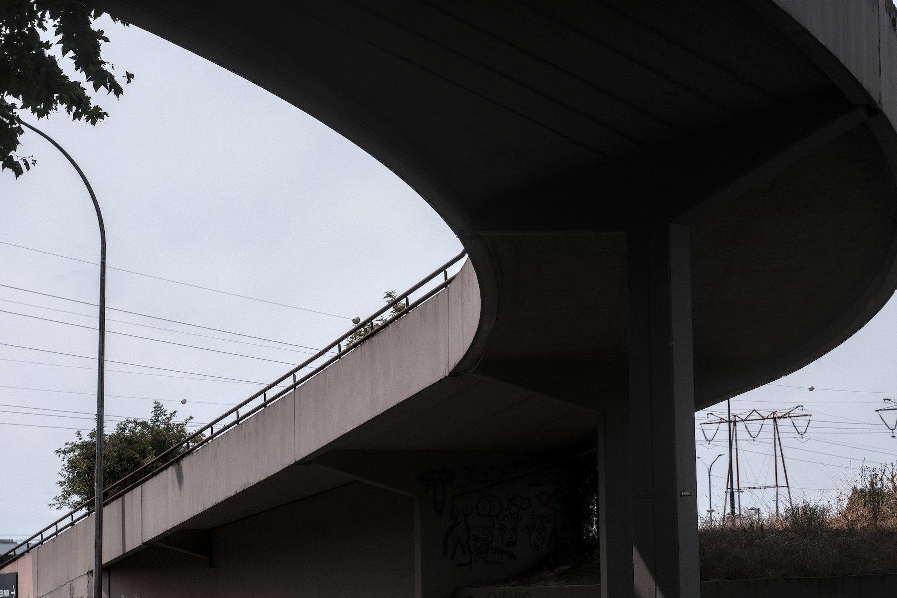 Photo horizontale. Vue de dessous d’une rampe routière qui tourne sur la gauche en descendant d’un niveau, sous un ciel gris