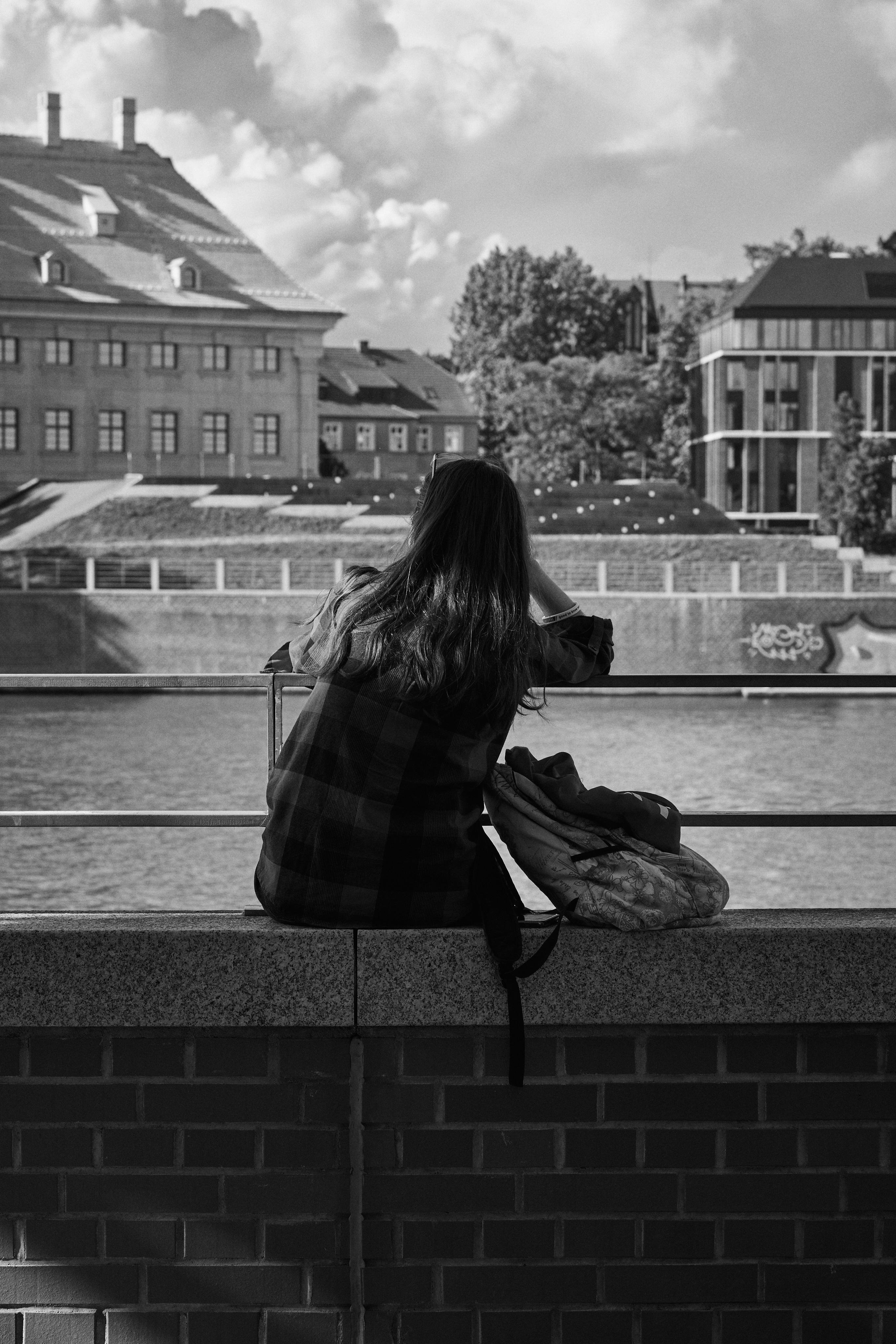 The image is in black and white and shows a person sitting on a ledge, facing away from the camera. The person has long hair and is wearing a checkered shirt. Next to them is a backpack. They are overlooking a body of water, possibly a river or canal. In the background, there are buildings, some of which have multiple windows and distinct architectural styles. Trees and clouds are also visible in the background. The overall scene appears calm and contemplative.
