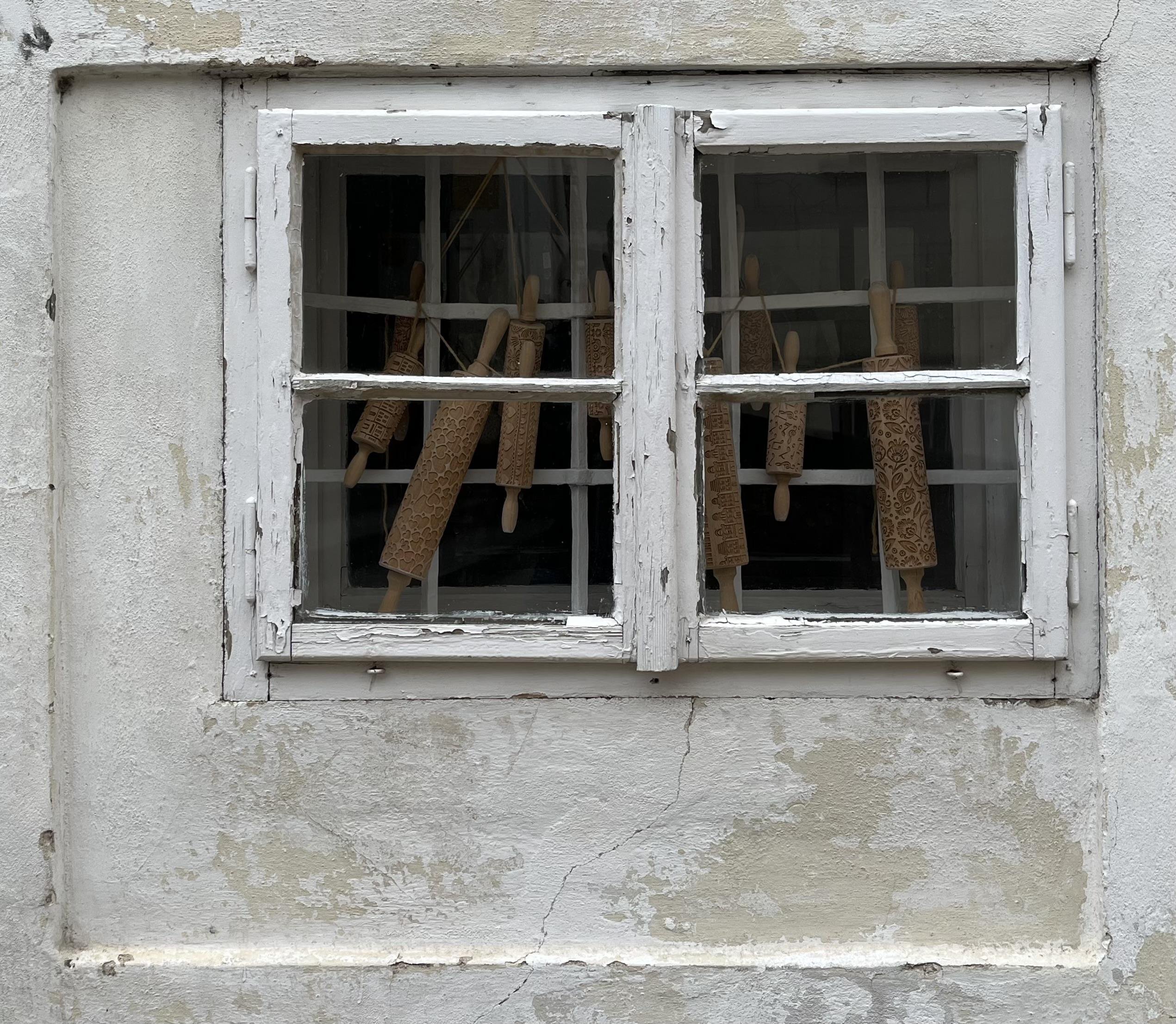 Weathered white-framed double window asymmetrically set in a cracked, peeling plaster wall, with several patterned wooden rolling pins visible behind the window panes and bars.