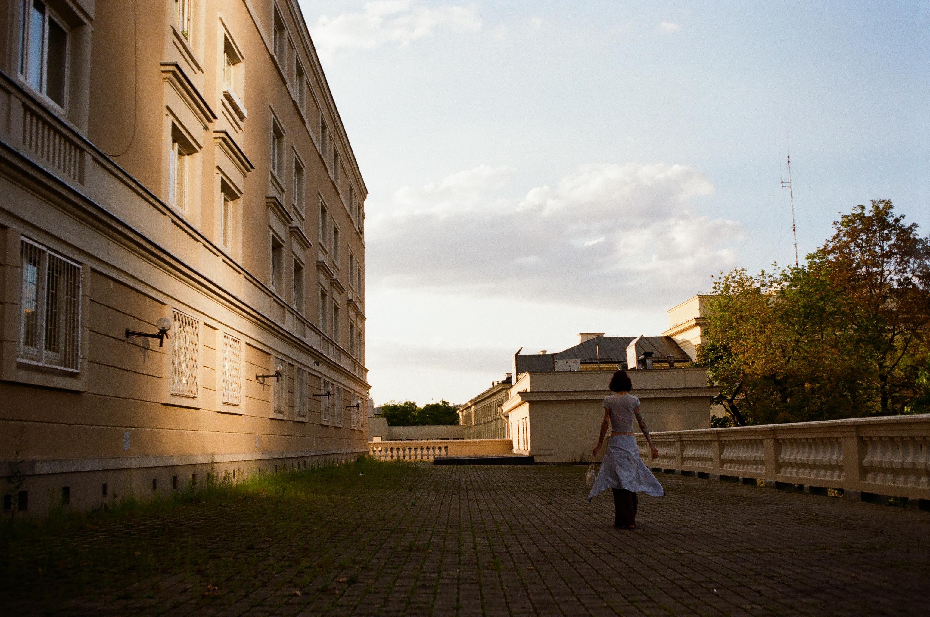 Woman in a light dress walking away along a cobblestone path beside a large beige building, with a white balustrade on the right and trees and low rooftops under a partly cloudy evening sky.