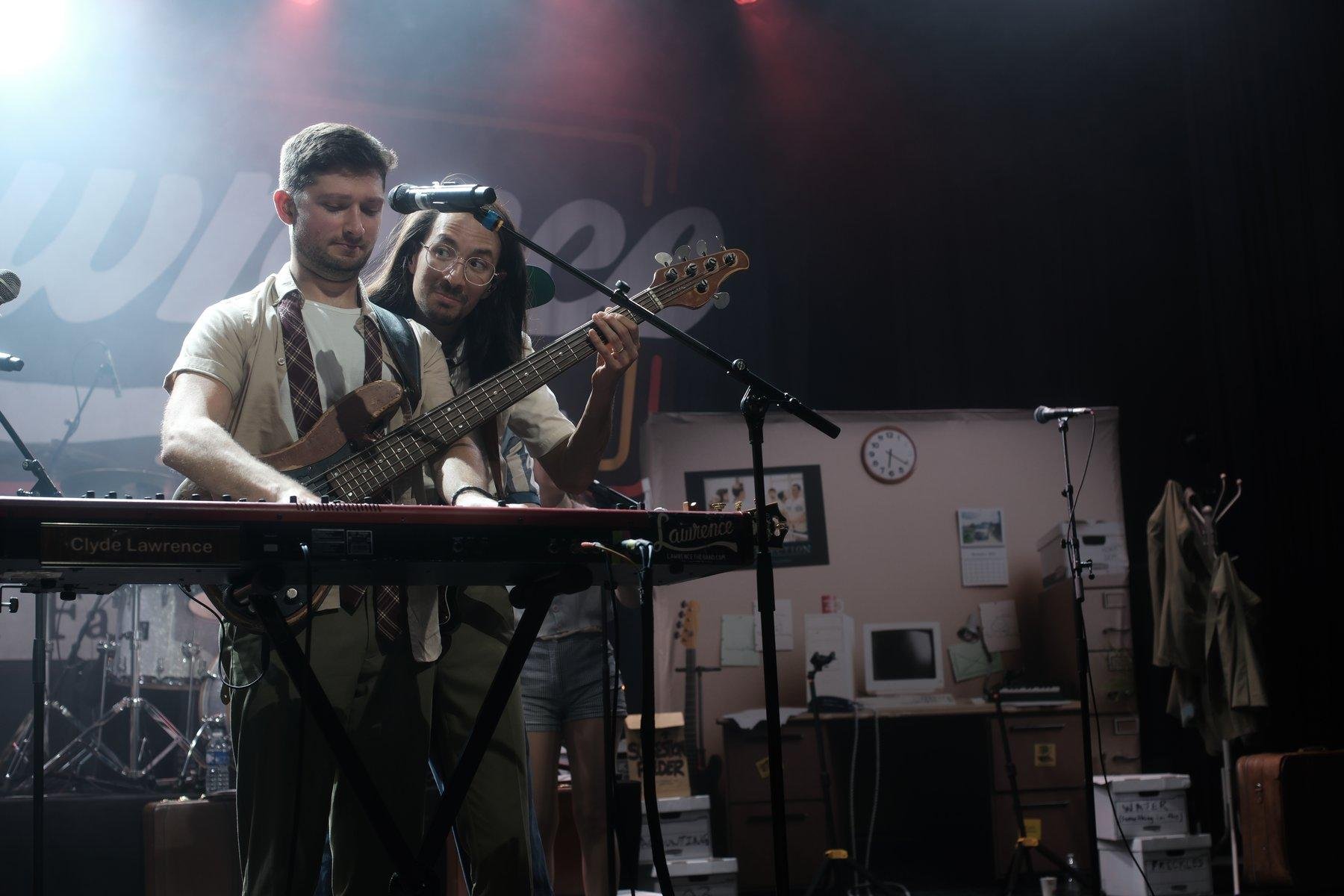 Photo horizontale. Un musicien joue du clavier en portant une basse qui est jouée par un collègue derrière lui et qui le regarde avec un air mi amusé, mi questionant