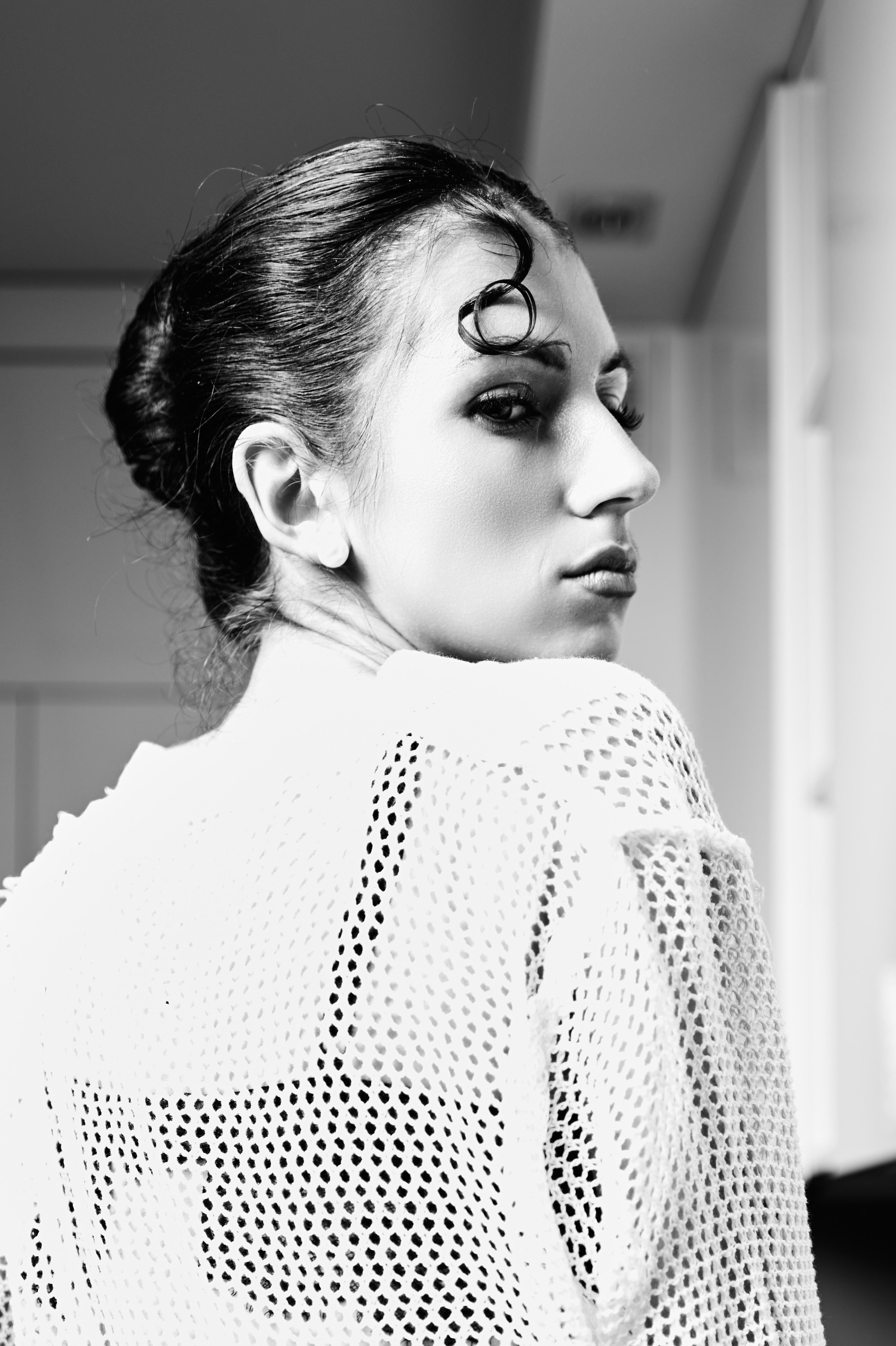 Black-and-white close-up portrait of a woman in profile, looking over her shoulder, with hair in a bun and a curled strand on her forehead, wearing a white mesh knit sweater against a softly blurred indoor background.