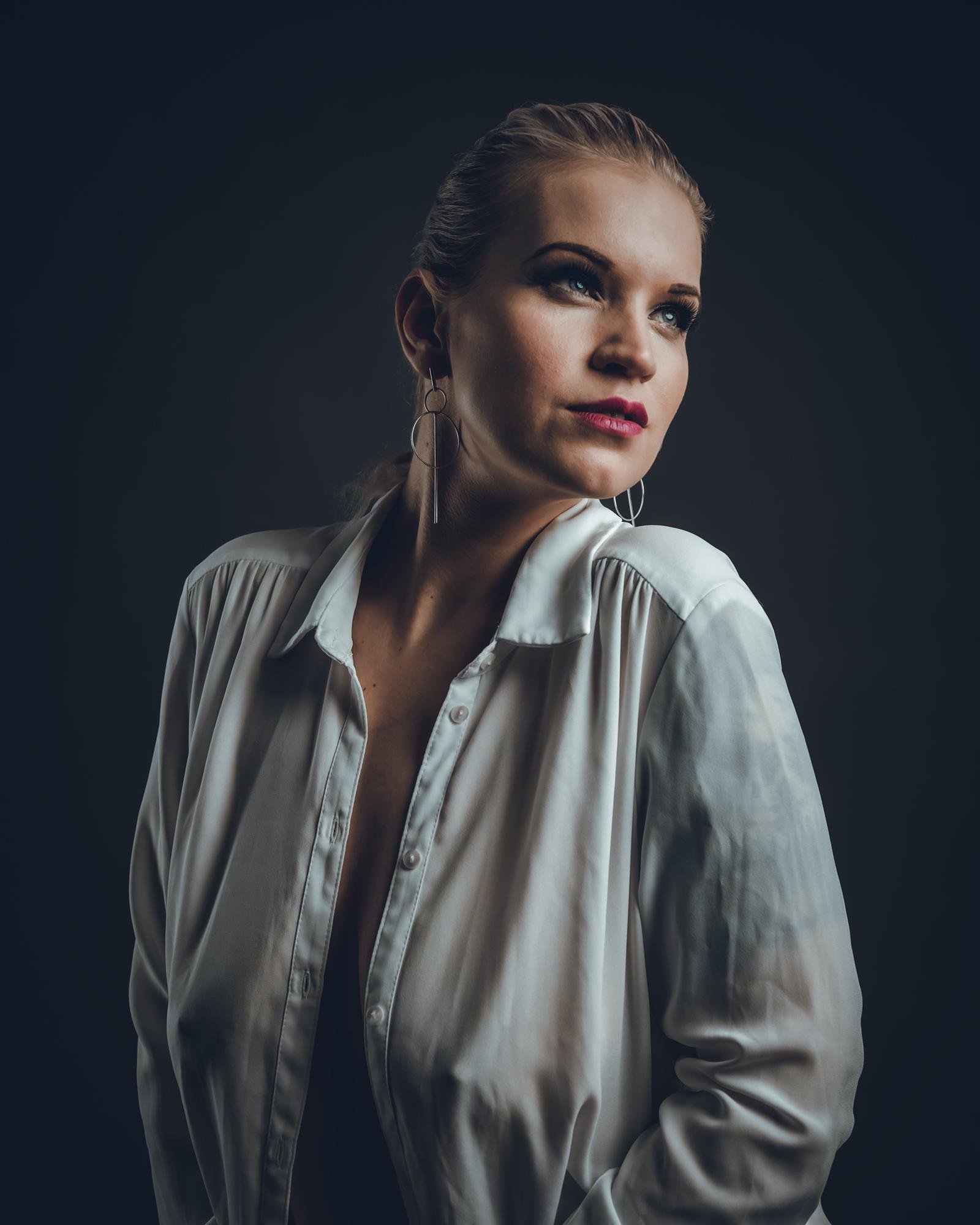 The image shows a person in a white shirt with an open collar, posing against a dark background. They have earrings and are looking slightly to the side. The lighting is dramatic, highlighting the features and creating shadows. Studiokuvaus Kuvaajankulma Lappeenranta
