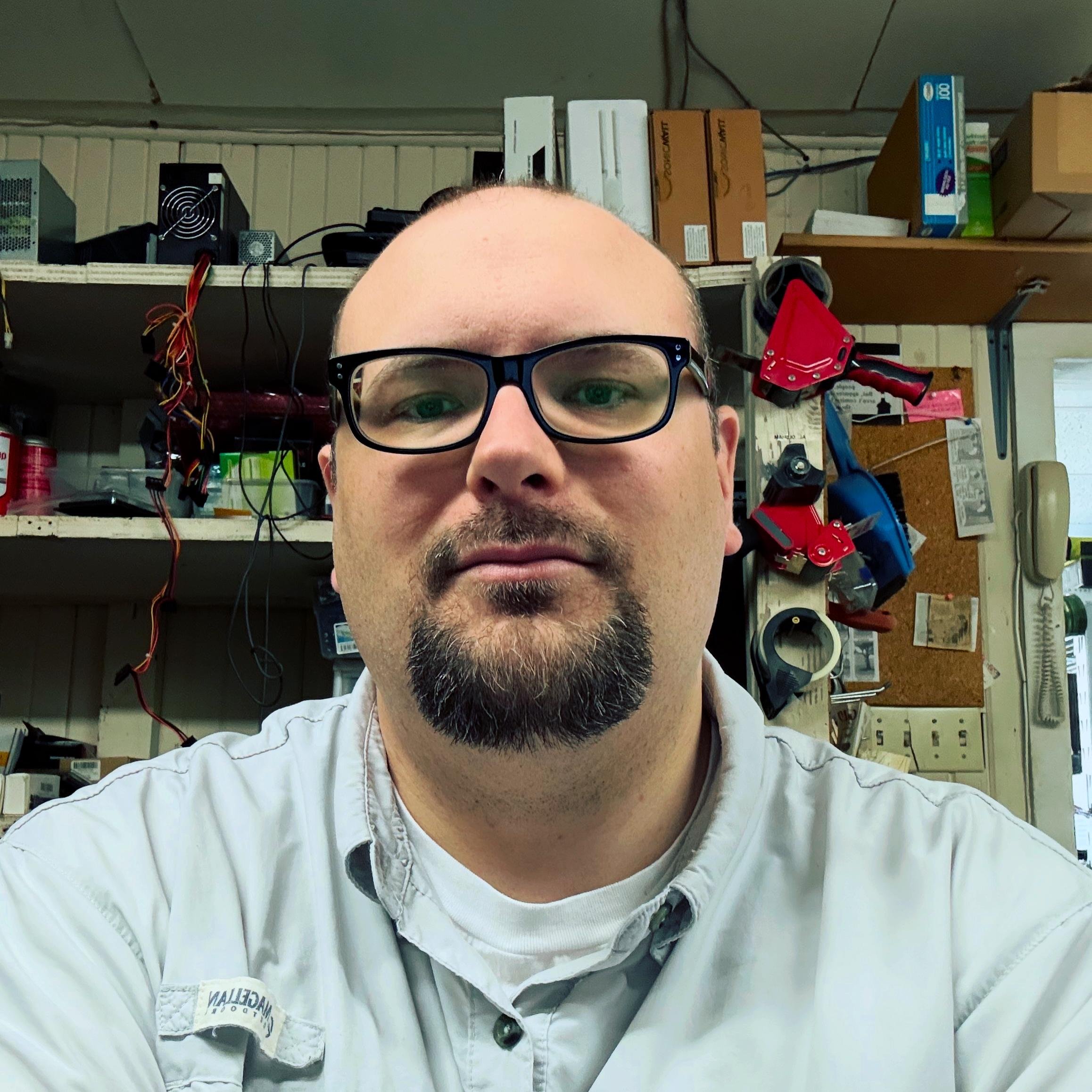 Photo of me wearing black-rimmed glasses and a light gray shirt sitting in a cluttered workspace. Shelves behind me contain various electronics, cables, and boxes. A wall-mounted corkboard displays papers, and a phone is visible on the right. The room has industrial and professional tools, including red tape dispensers.