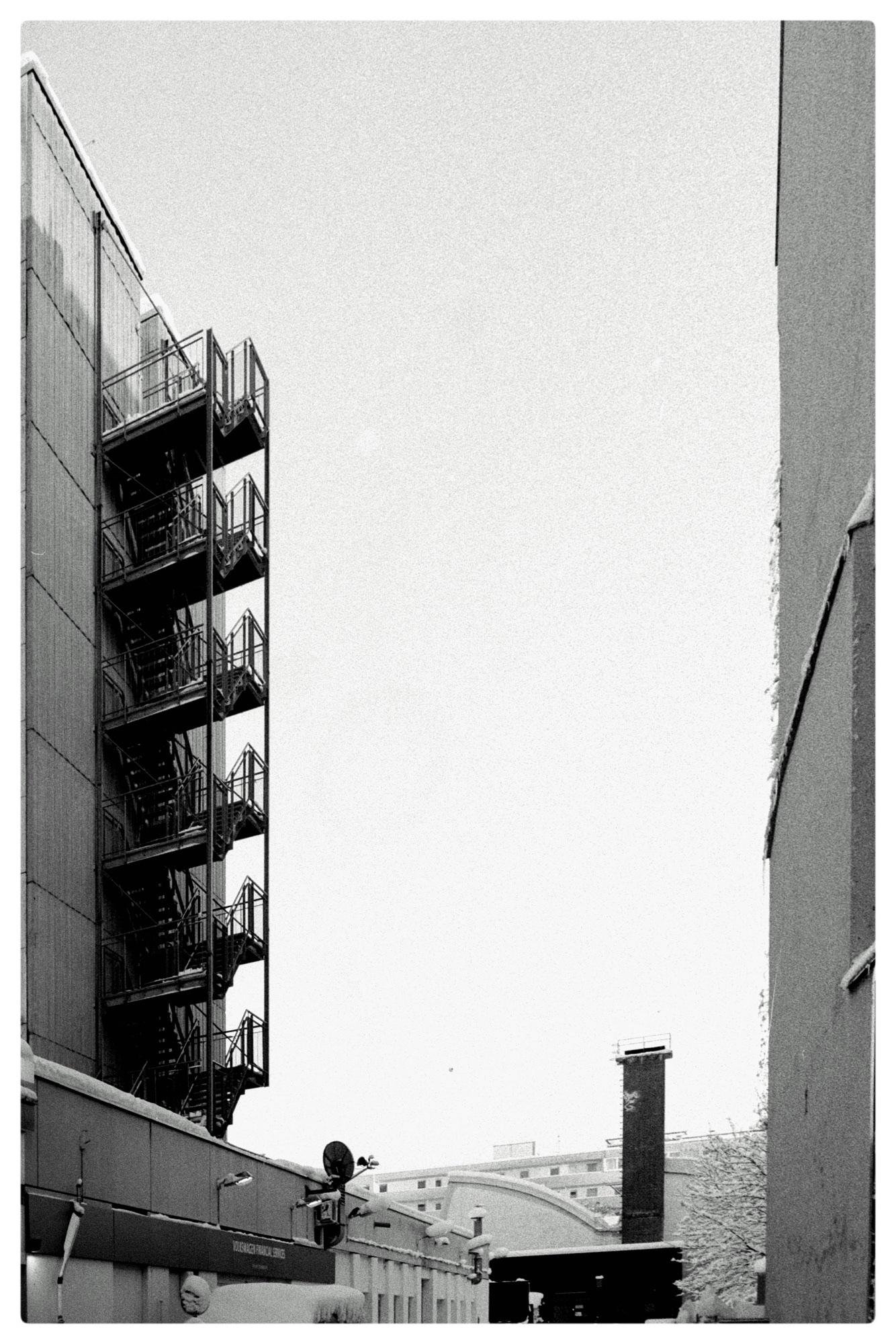 Black-and-white view down a narrow alley between two tall buildings, with an exterior metal fire-escape staircase on the left, a wide pale sky above, and a small industrial yard at the bottom featuring a satellite dish, a tall chimney stack, and snow-covered surfaces.