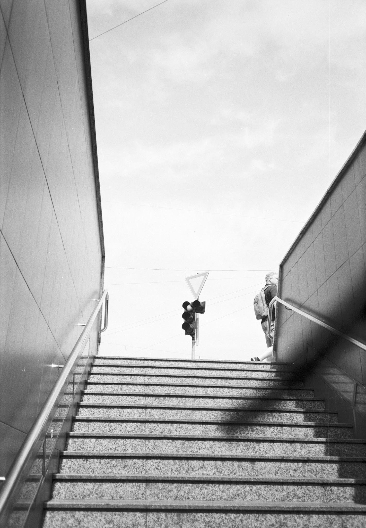 Black-and-white view from the bottom of a stairway between two tiled walls with metal handrails, leading up to an outdoor opening with sky, a traffic light and triangular yield sign, and a person with a backpack near the top right.