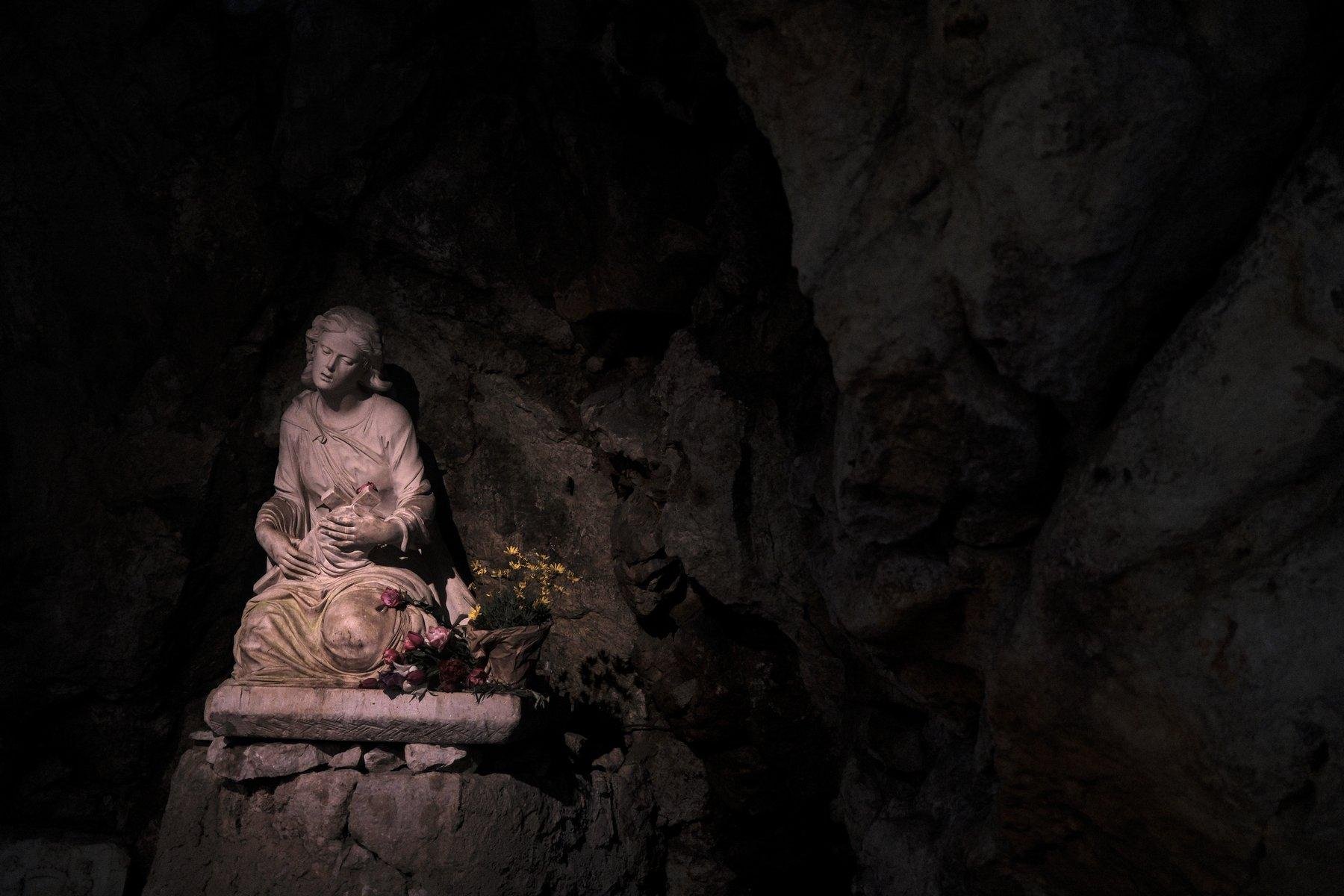 Photo horizontale. Dans une grotte sombre, une statue de (je crois) Marie-Madelaine, qui tiens une croix sur laquette est collée une rose. Son visage semble exprimer la souffrance. Des fleurs sont posée à droite. La statue est la seule chose bien éclairée de la scène, mais on devine les parois de roches