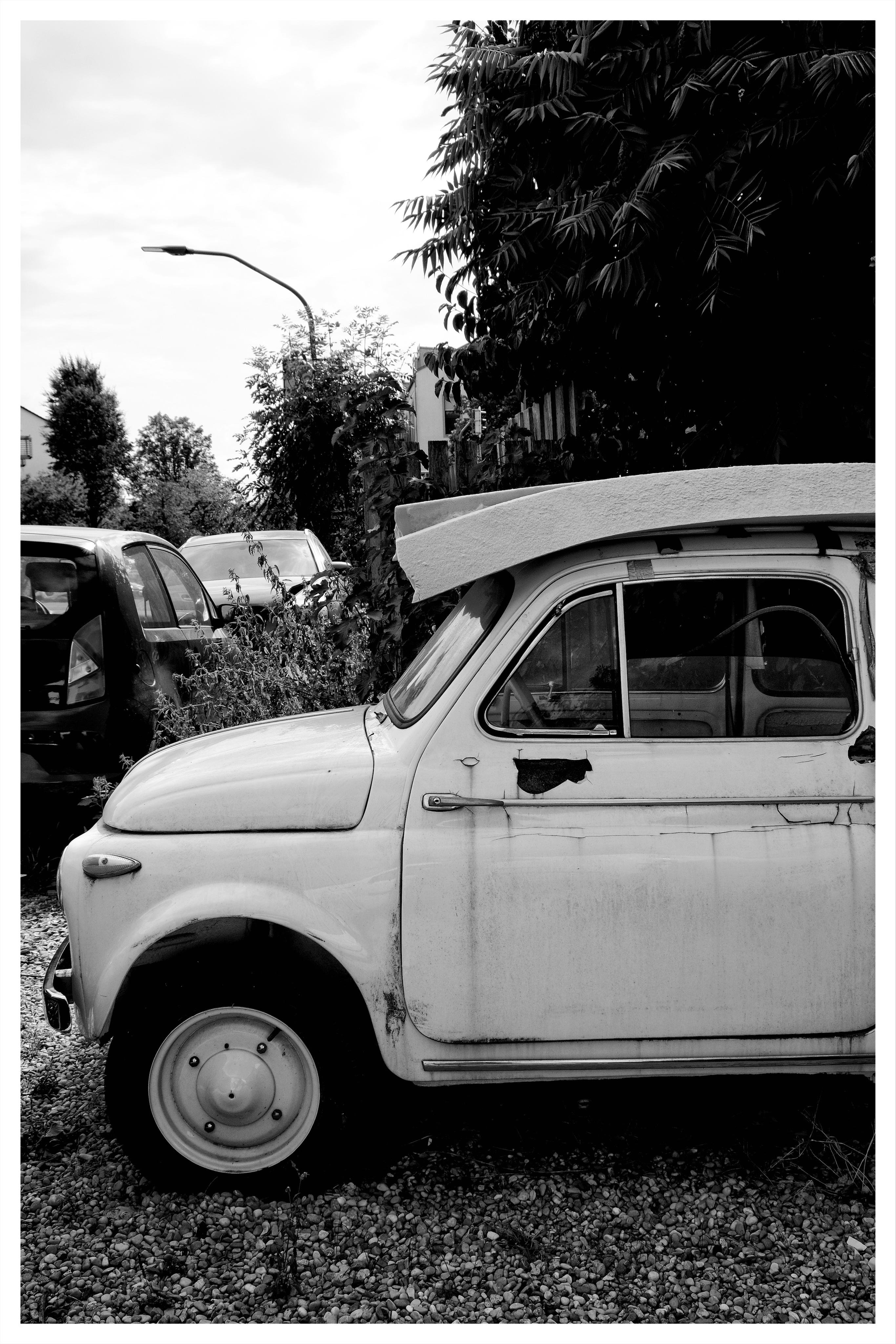Black-and-white photo of an old compact car parked on gravel, shown from the side with the front wheel and door in the foreground; other parked cars, a streetlamp, and dense trees line the background.