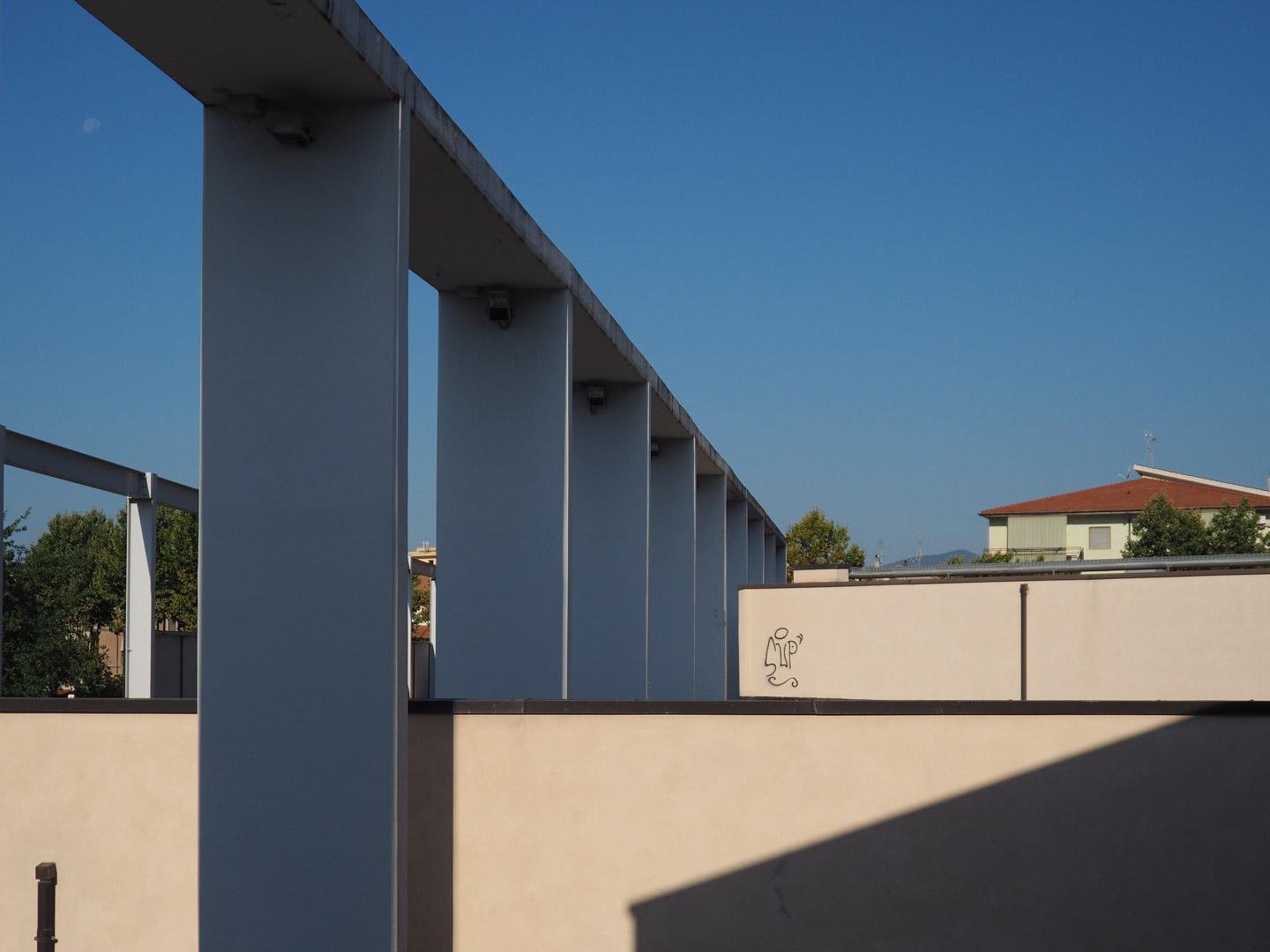 Strutture in cemento e marmo dallo scopo prevalentemente ornamentale, composte da pilastri squadrati di cemento su cui sono posti dei blocchi orizzontali sempre di cemento, che conferiscono all'opera la sembianza di un ponte. Un altra struttura simile, a sinistra, è realizzata con travi di verro. In basso, pareti verticali beige illuminate dal sole, che vi disegna anche alcune ombre. A destra è visibile l'ultimo piano di un'abitazione, con relativo tetto spiovente. Il cielo è limpido e azzurro.