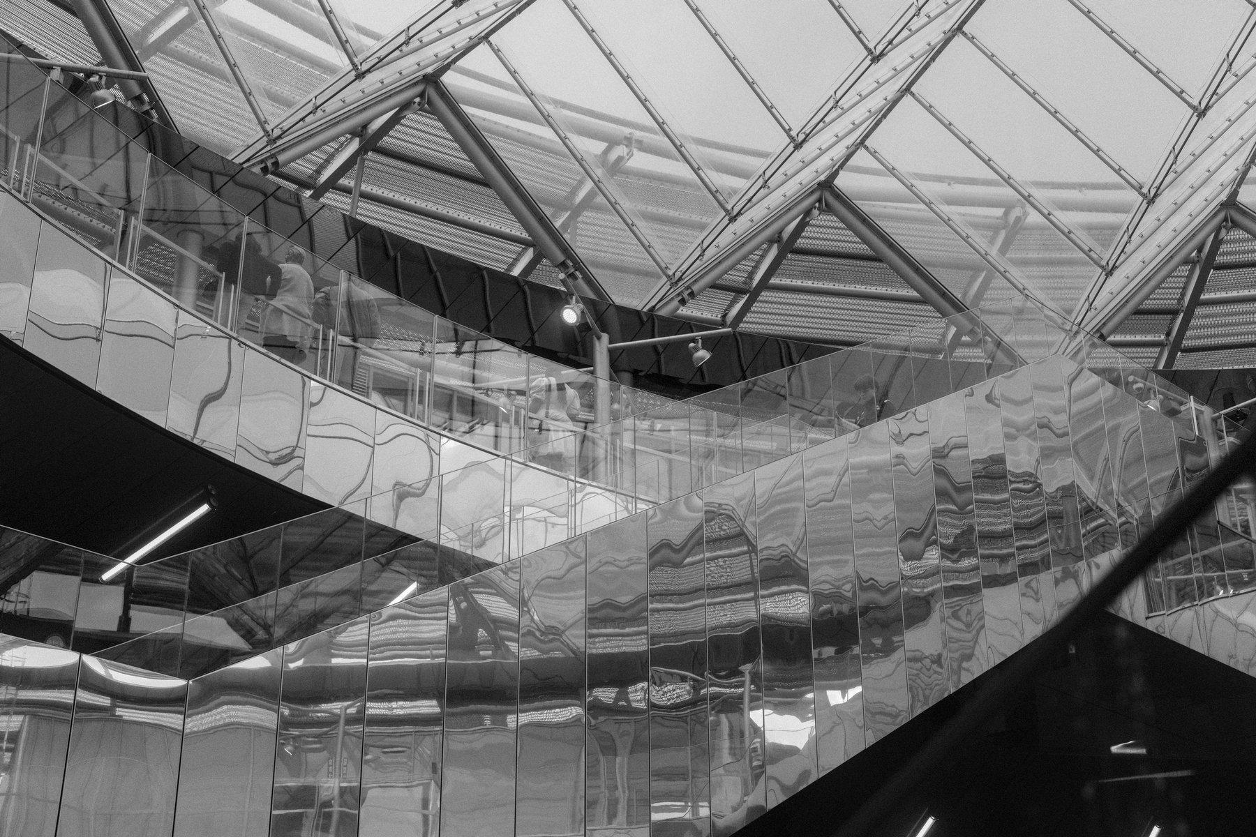 Photo horizontale en noir et blanc. Une rampe d’escalator aux parois en métal réfléchissant, une passerelle avec ses vitres de sécurité en plexiglass et un bout de la verrière de la station Villejuif Gustave Roussy