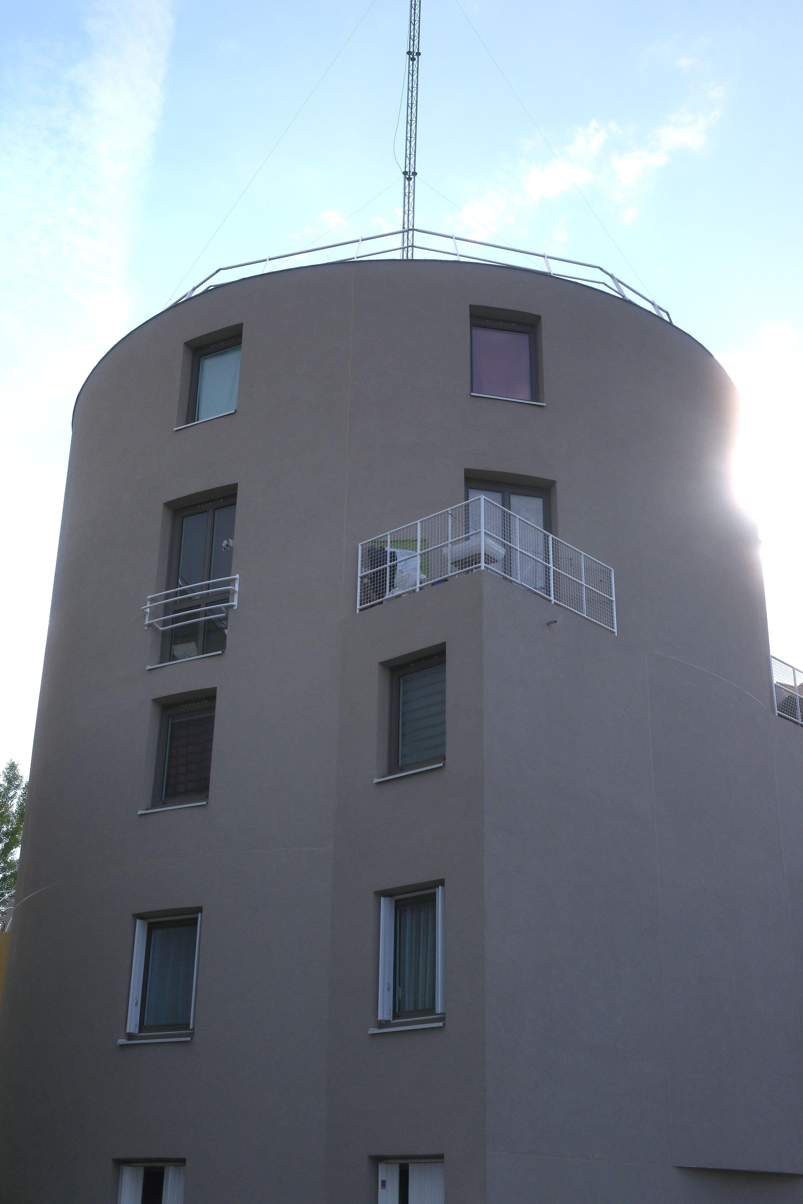 A modern gray building which is round but with a square part into it giving the impression of a change of mind.  There’s an antenna on the roof, set against a partly cloudy sky.