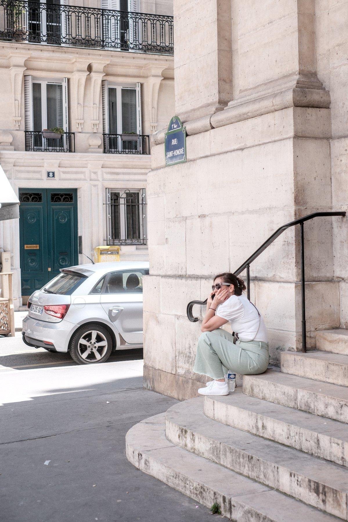 Photo verticale. Dans la rue, une jeune femme brune au cheveux attachés par une pince, lunettes de soleil, haut blanc, pantalon vert pale, baskets blanches. Elle est assise sur le coin d’une marche d’escalier en pierre, une petite bouteille d’eau entre ses pieds, en train de parler au téléphone
