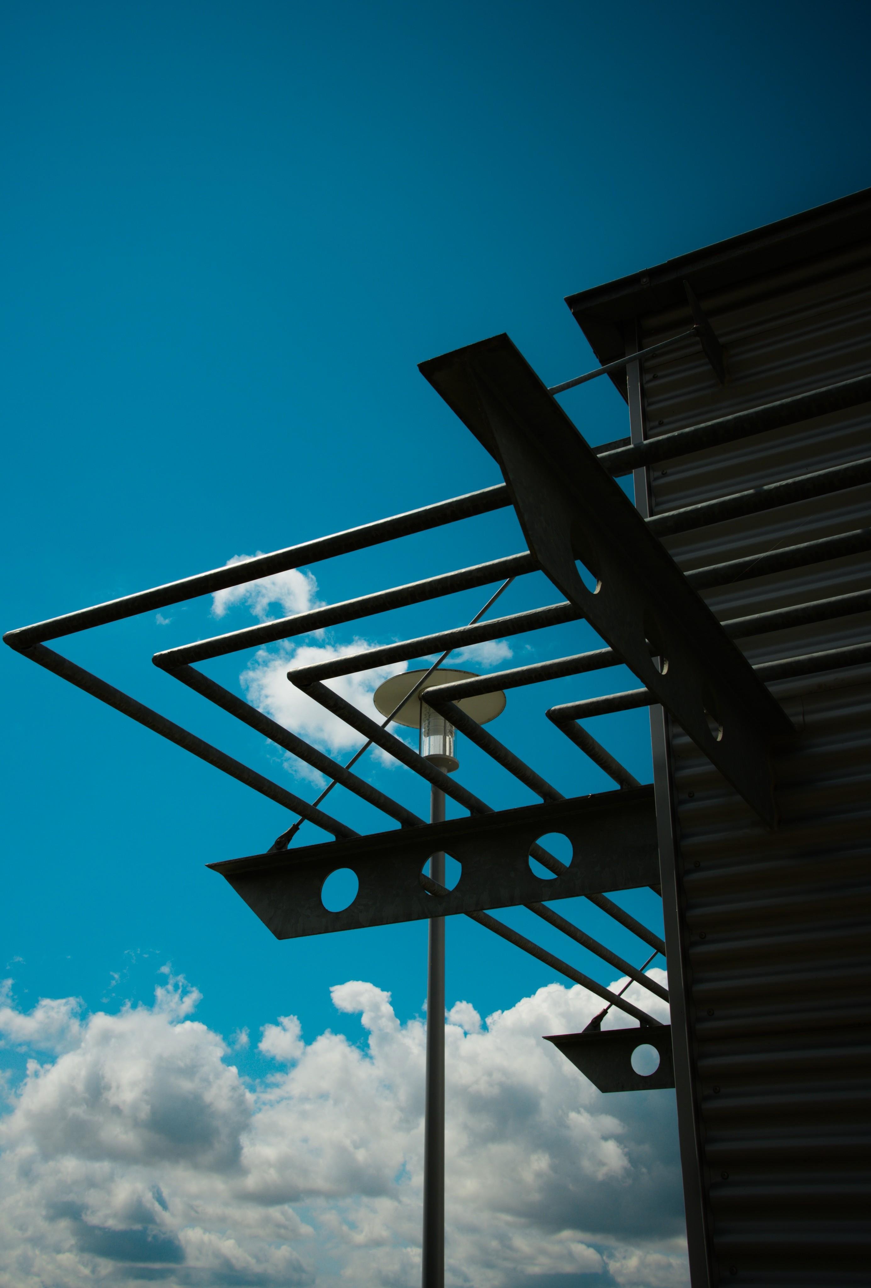 Upward view of a building corner with dark metal awnings and horizontal beams, a streetlight pole in the center behind it, and a bright blue sky with scattered white clouds in the background.