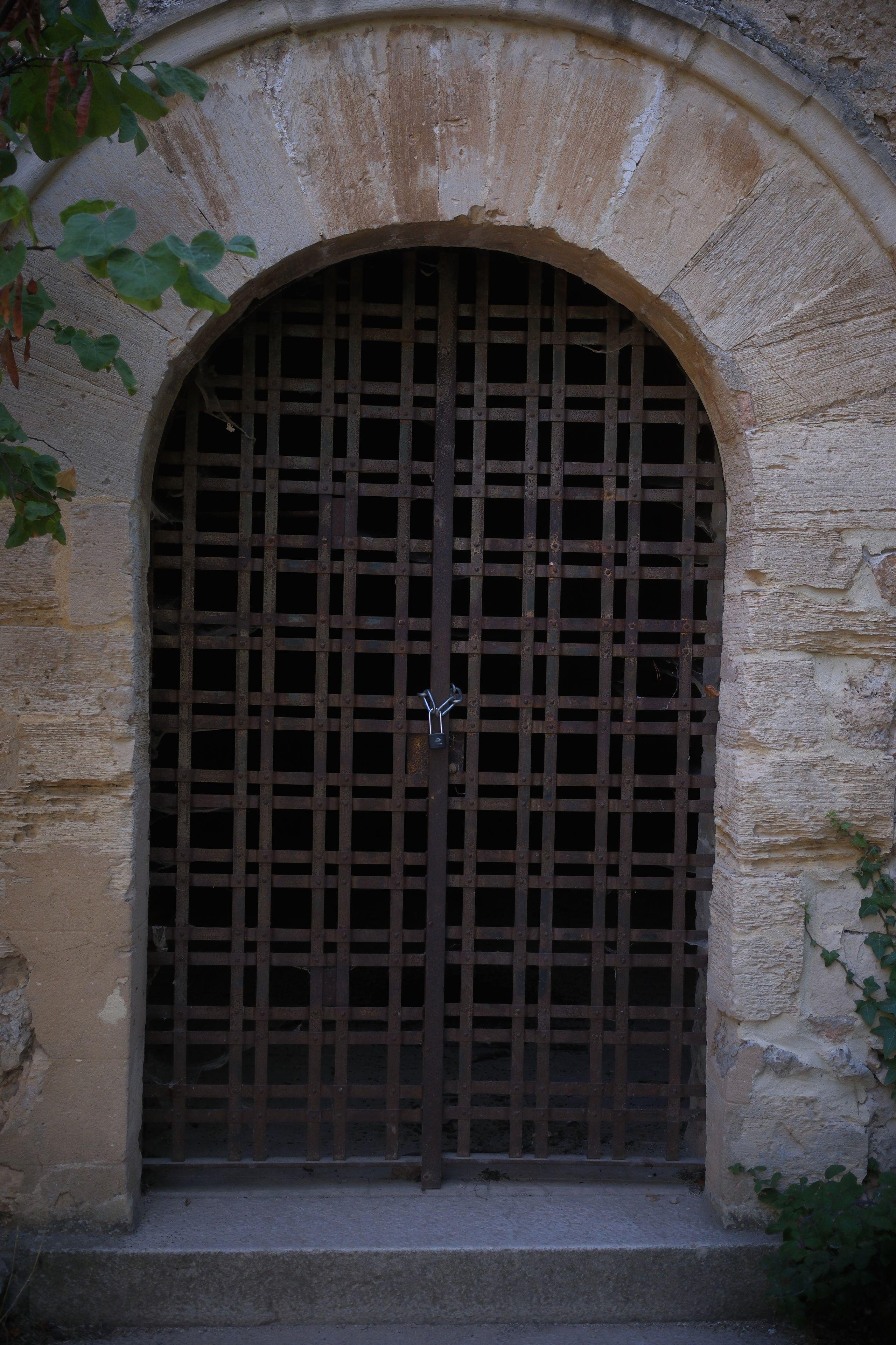Photo verticale. Une vieille porte faite en grille métallique rouillée et tenue fermée par une chaine et un cadenas