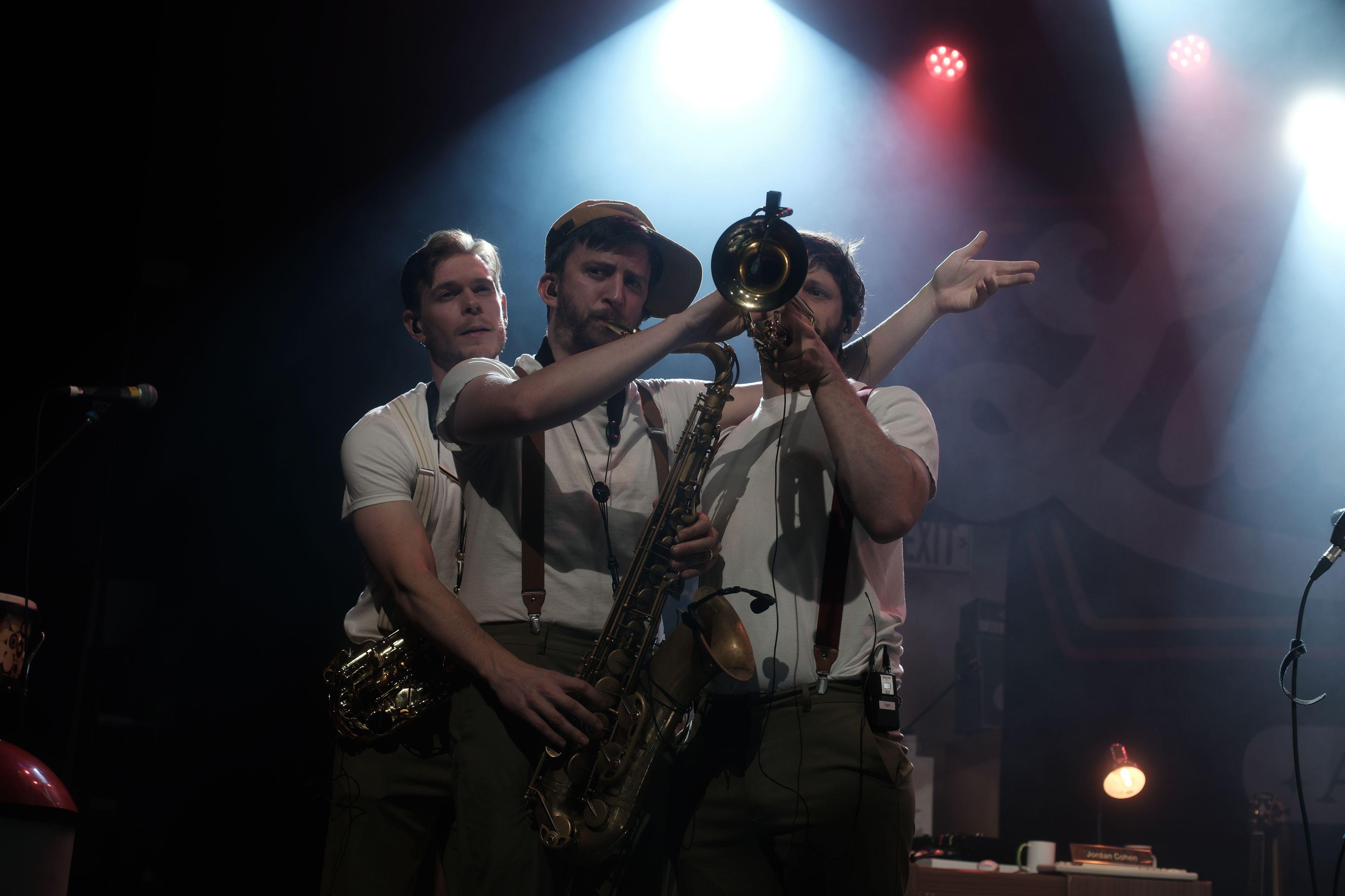 Photo horizontale. 3 musiciens sur scènes, collés les uns aux autres, celui de droite soufflant dans une trompette, mais qui est jouée par son comparse du milieu, qui souffle dans son saxophone, joué par les mains du comparse de gauche