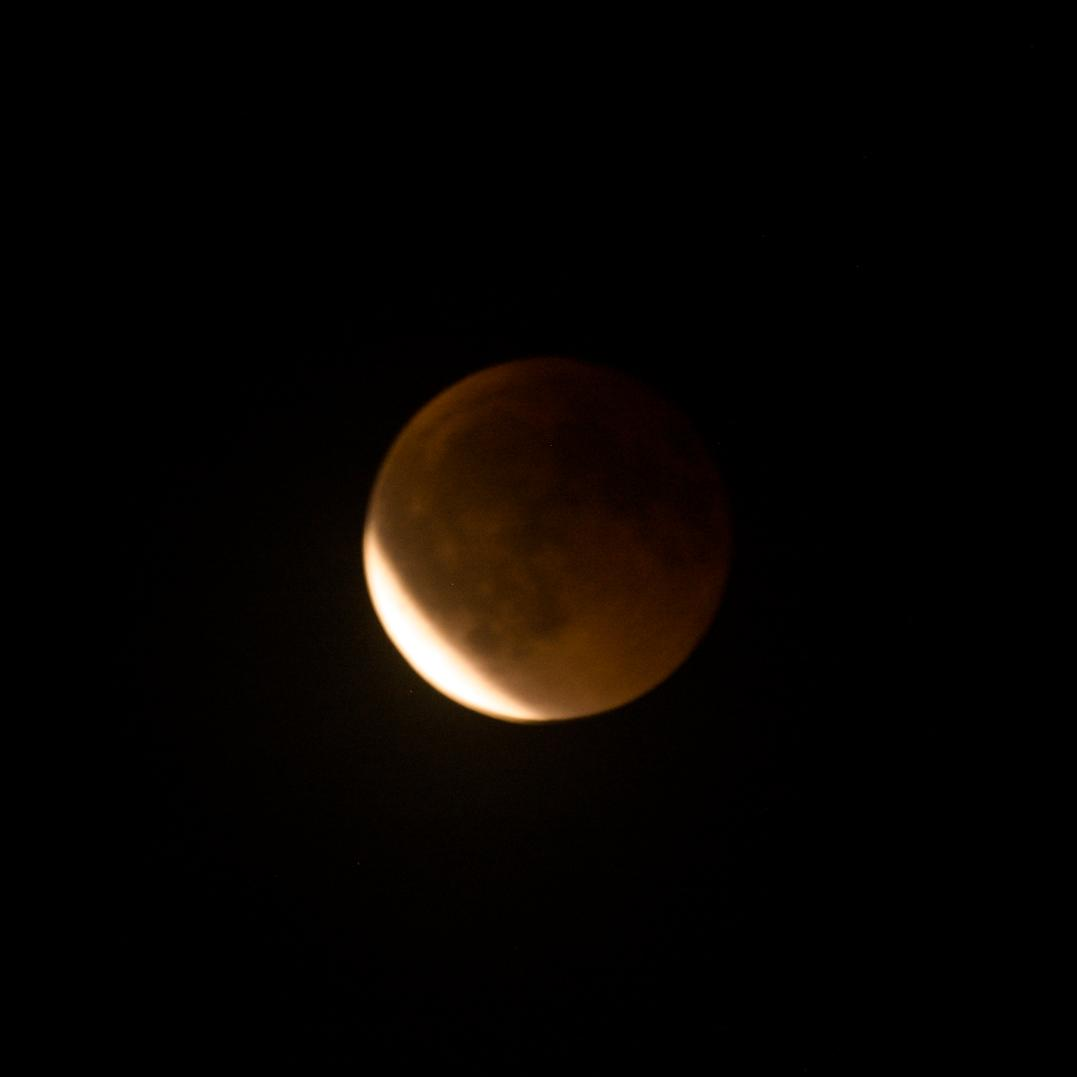 Lunar eclipse with the moon partially obscured, casting a reddish-brown hue across its surface. The left side of the moon remains visible with a bright, curved edge against a dark night sky.