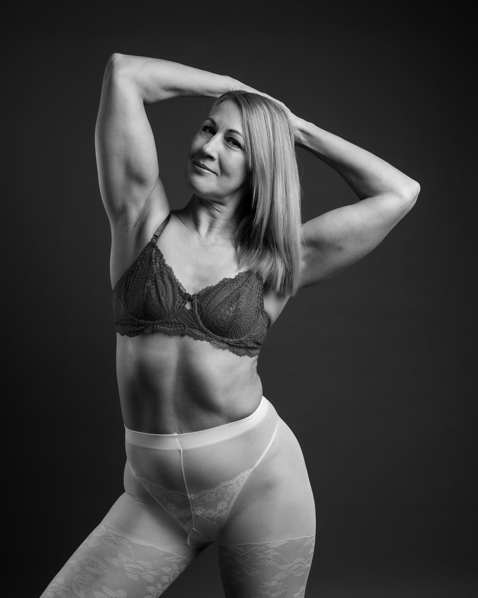 The image shows a woman posing in underwear, consisting of a bra and sheer tights. It's presented in black and white.