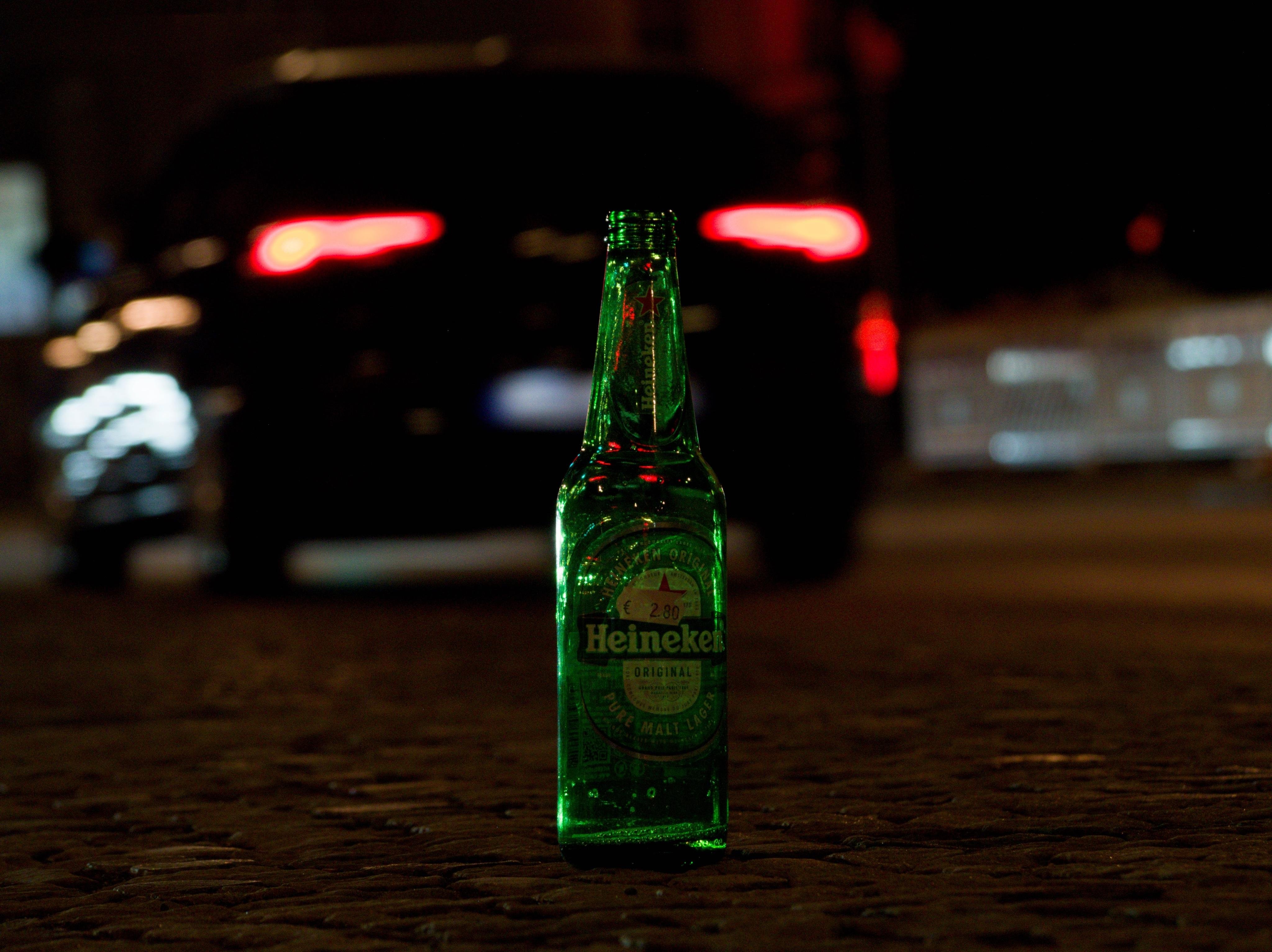 A green Heineken beer bottle on a dark surface, with a dark car and bright red taillights in the background.
