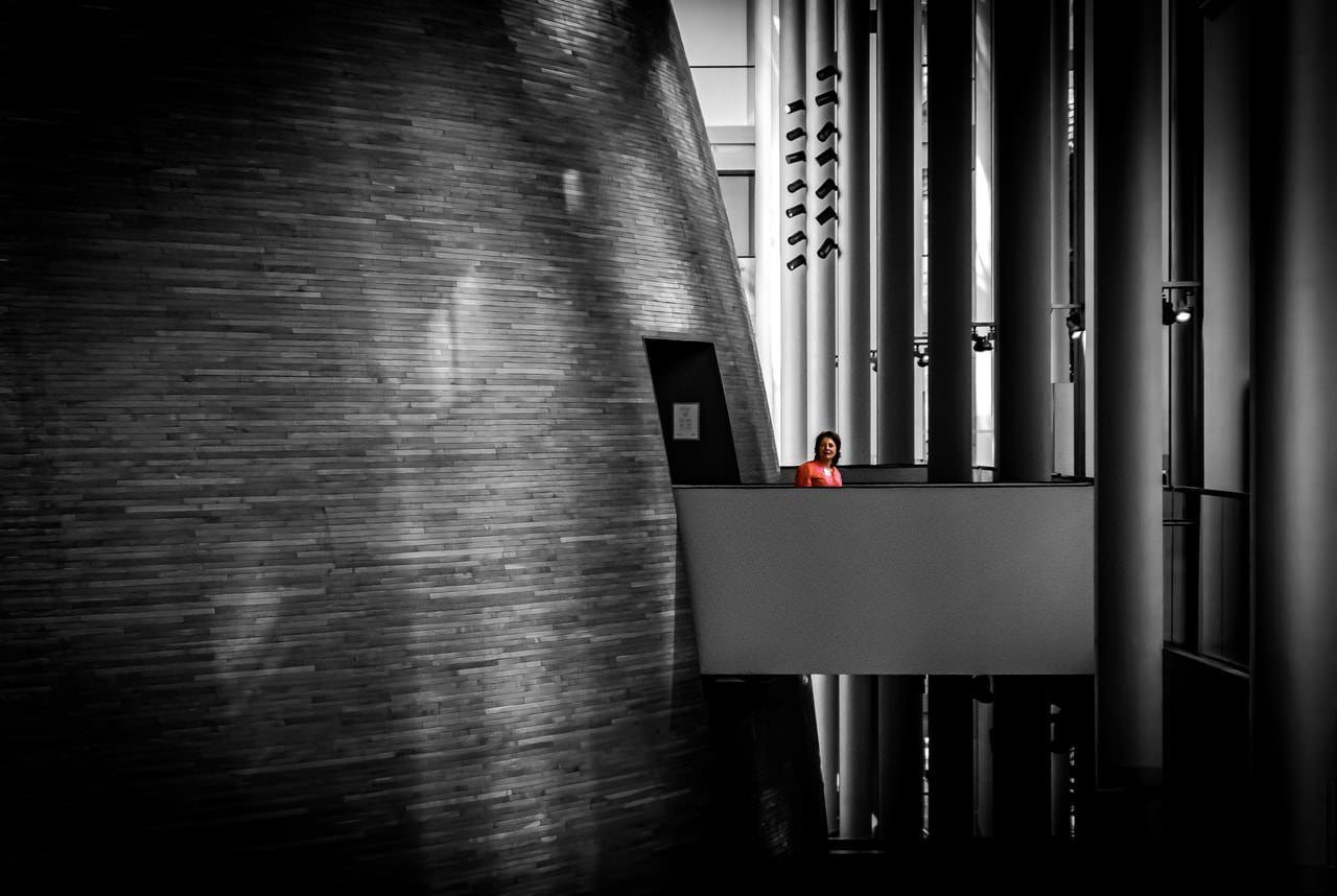 A person in a red top stands on a modern balcony inside a building. The background features a large, textured wall on the left and a series of vertical columns on the right, creating strong contrasts between light and shadow. The composition emphasizes geometric shapes and lines.