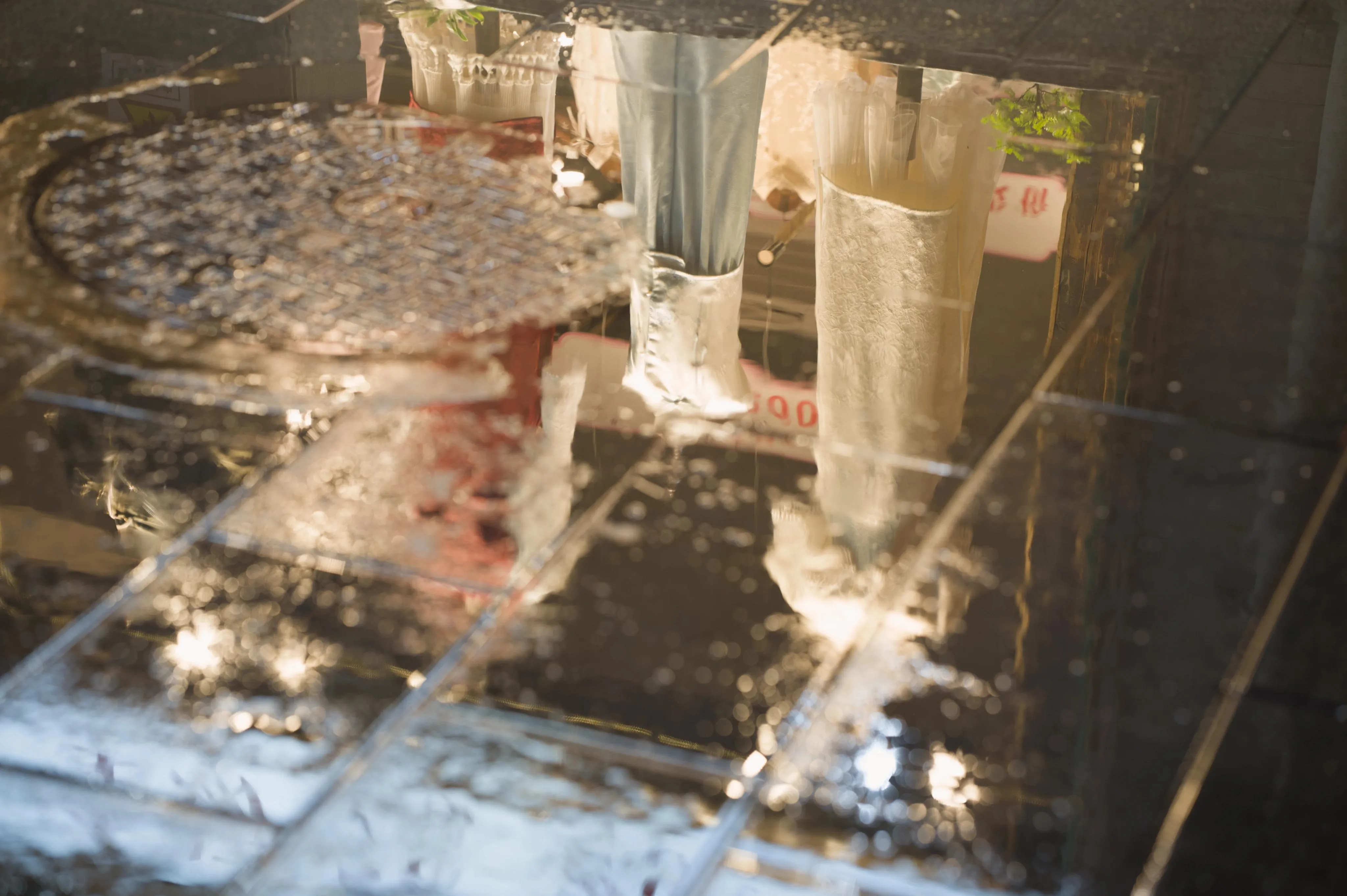 Photograph of reflections of dresses in a store store window on the wet street pavement. A sewer grate obstructs part of the dresses.