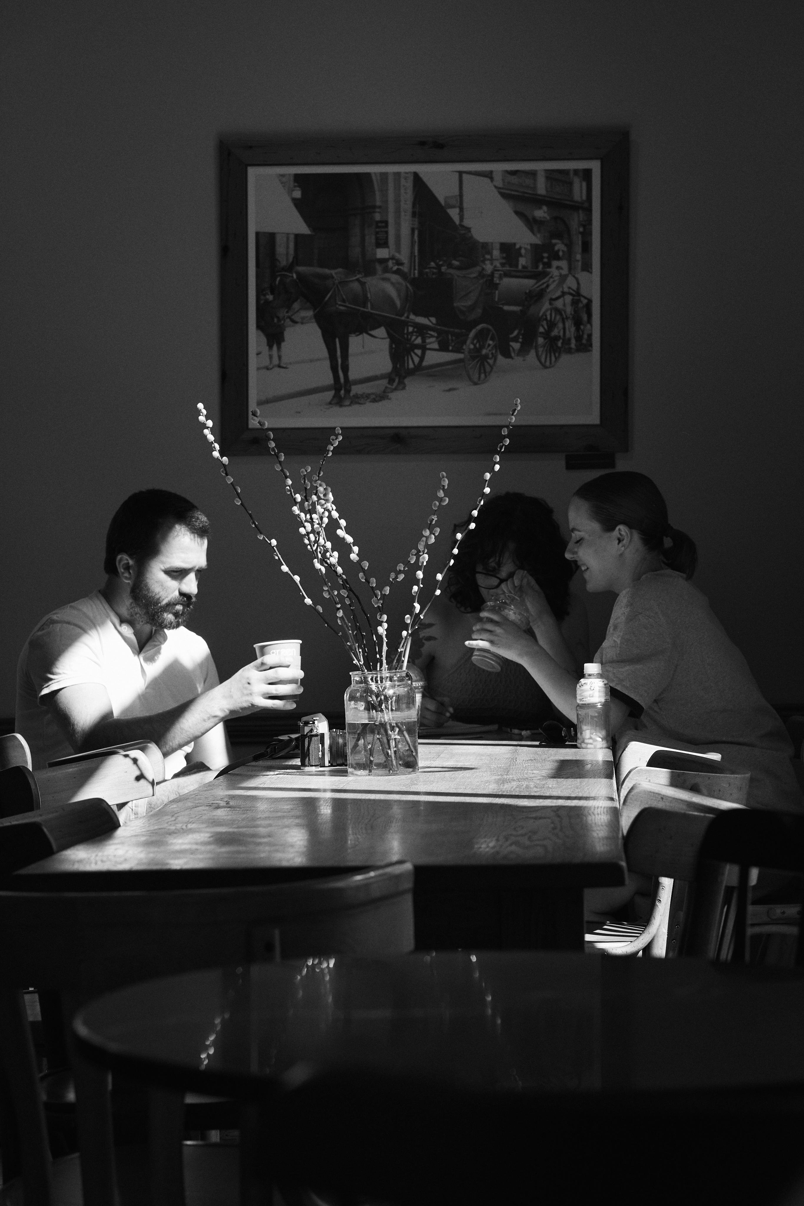 Black-and-white photo of three adults seated at a wooden table in a café, each holding a drink; a glass jar with tall flowering branches sits in the center, and a framed picture of a horse-drawn carriage hangs on the wall behind them.