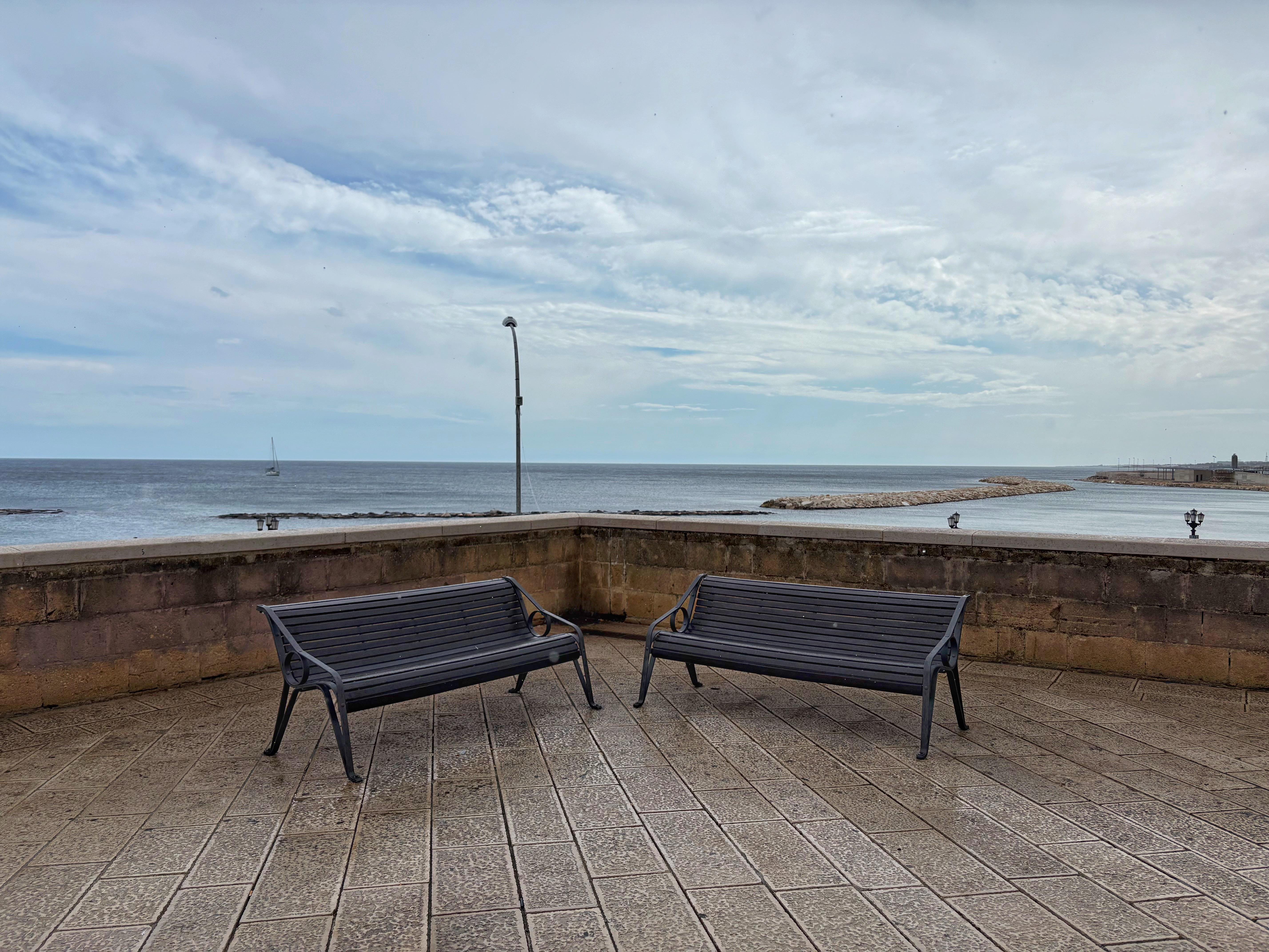 Two black metal benches on a paved terrace next to a stone wall overlooking the sea. A lamppost stands near the wall, with a sailboat visible on the horizon under a partly cloudy sky.
