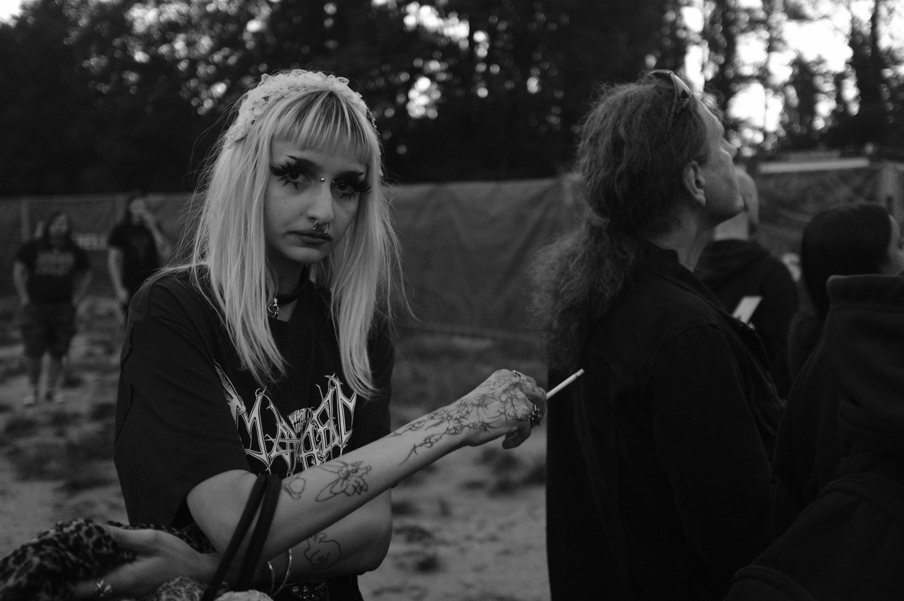 Young woman with long blonde hair and a decorative headband stands in an outdoor setting, holding a cigarette. She has dramatic eye makeup and facial piercings. Her arm adorned with tattoos is visible, and she wears a dark graphic t-shirt. Several people and blurred trees are in the background, captured in black and white.