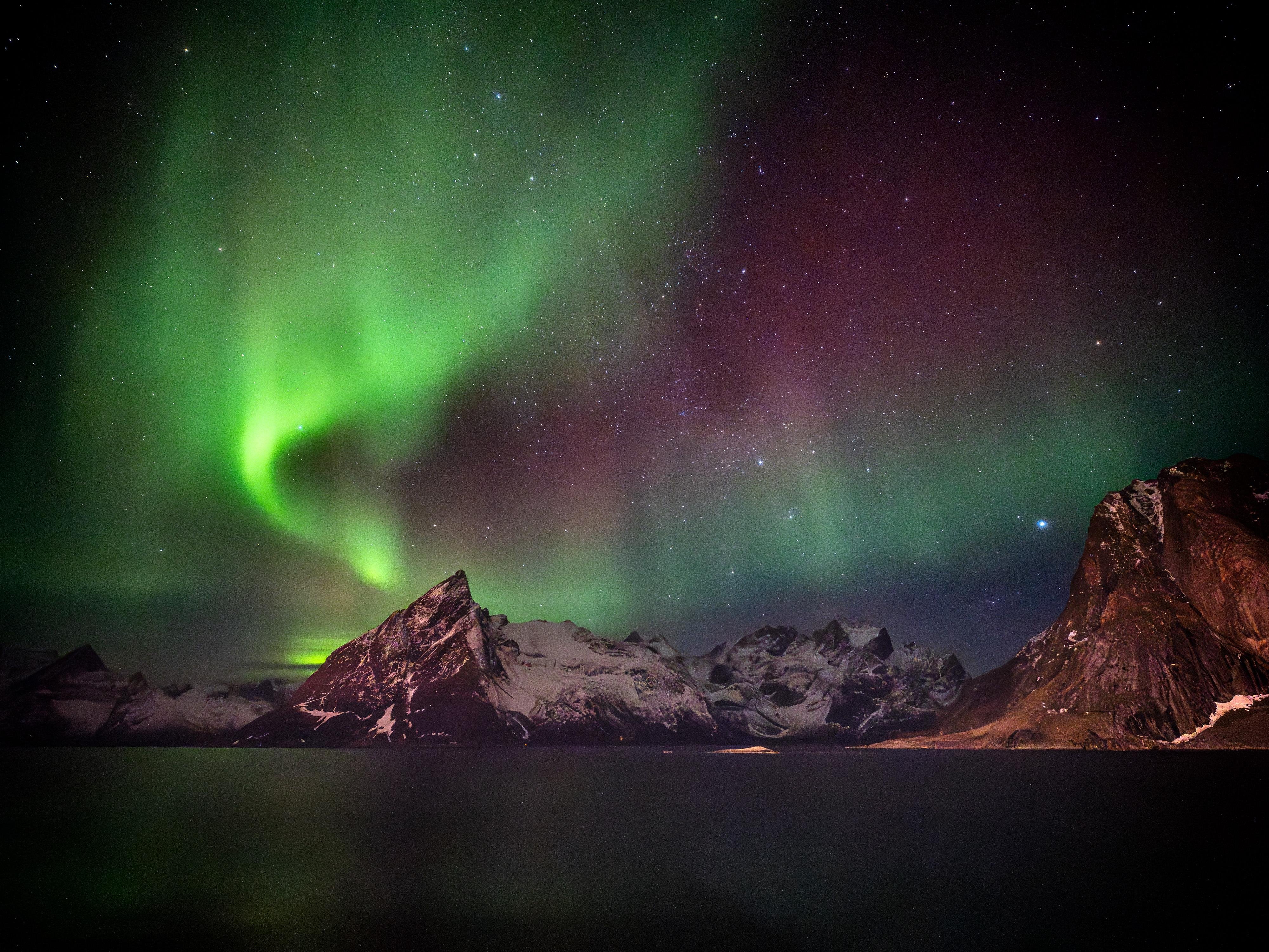 This image depicts the Northern Lights, or Aurora Borealis, illuminating the night sky with vibrant green and purple hues. The scene includes a rugged, snowy mountain landscape, possibly in a northern region with clear views of the auroras. The sky is also filled with stars, contributing to the stunning natural display.
