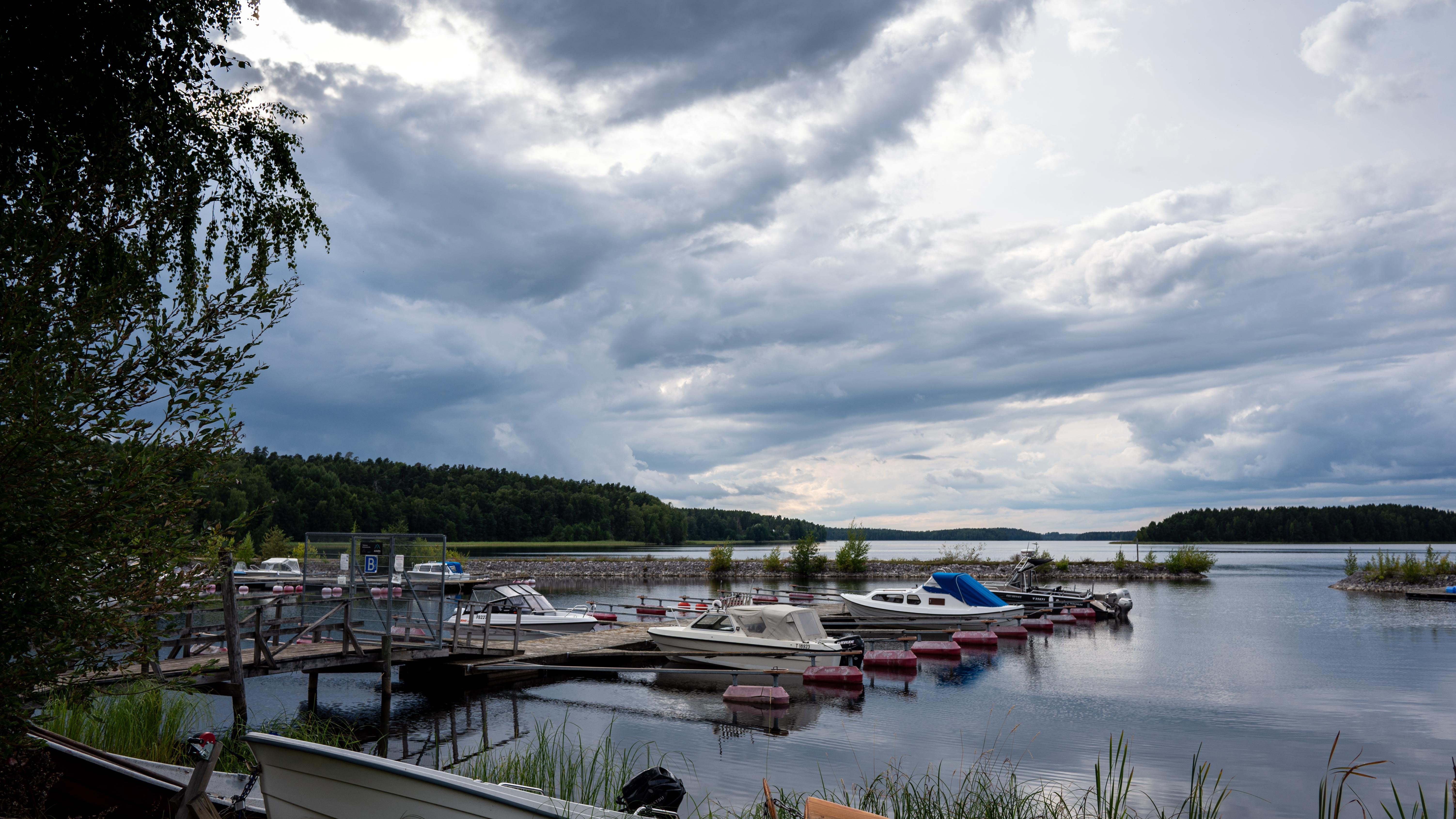 The image depicts a lakeside scene with several small boats docked at a wooden pier. The sky is cloudy, suggesting overcast weather, and the water is calm. There are trees and vegetation along the shoreline, contributing to a serene and natural atmosphere.