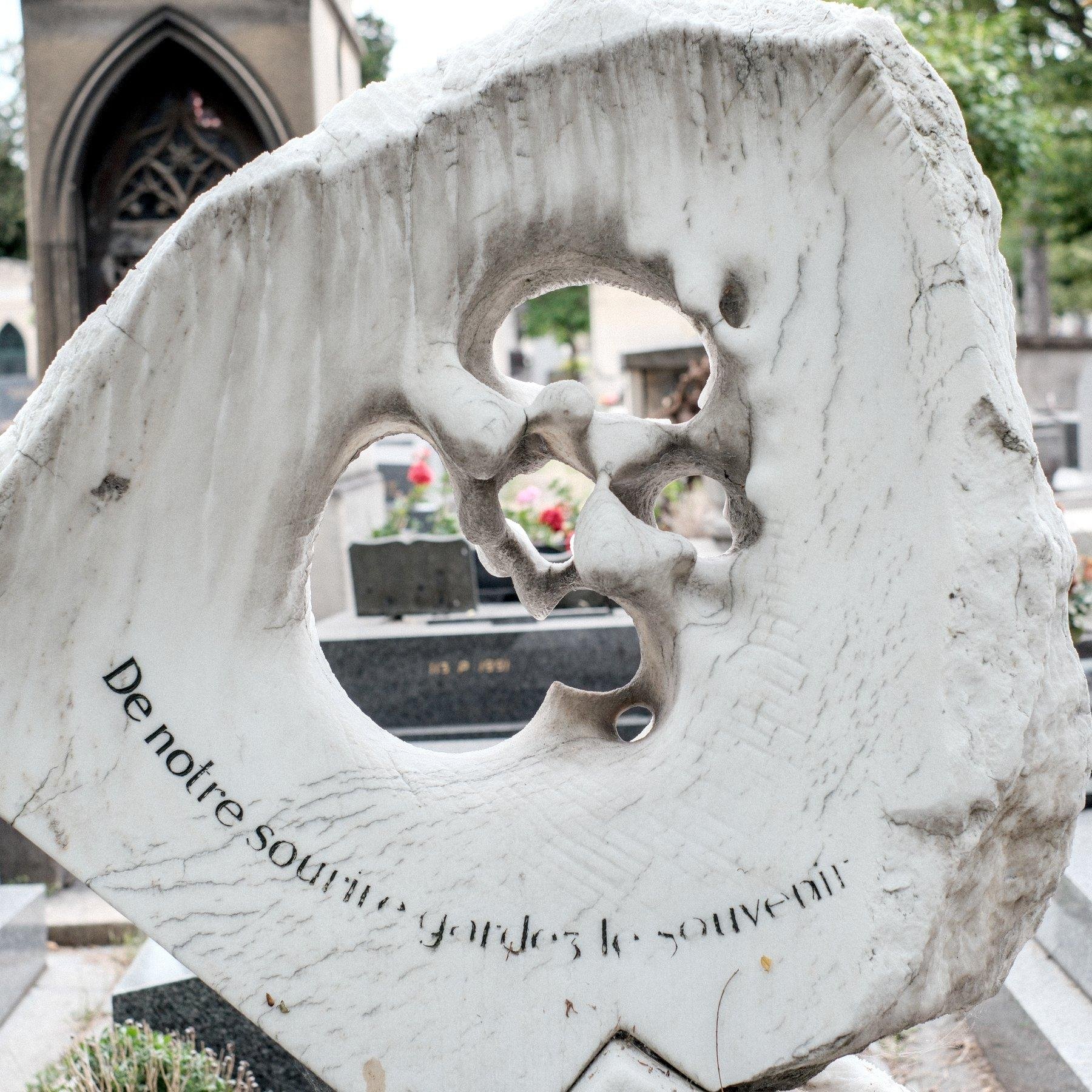 Photo carrée. Une stèle artistique en pierre (marbre blanc ?) sculptée avec des grands trous difformes au centre. Derrière, une fleur rouge dans le trou du centre