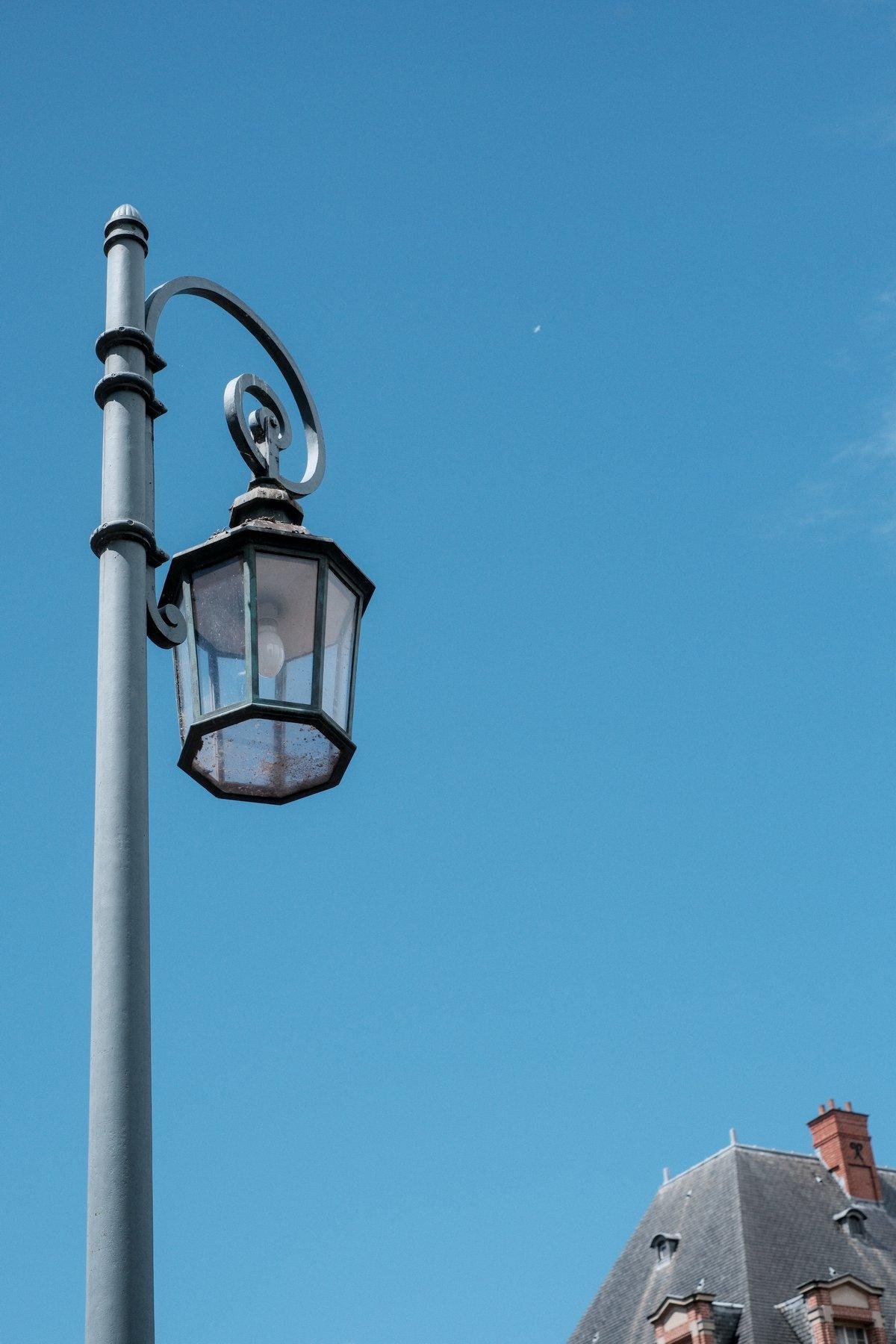 Photo verticale. À gauche contre plongée sur un lampadaire ouvragé sur un ciel bleu. Dans le coin inférieur droit, un bout de toit de bâtiment en ardoise et petite cheminée en brique rouge