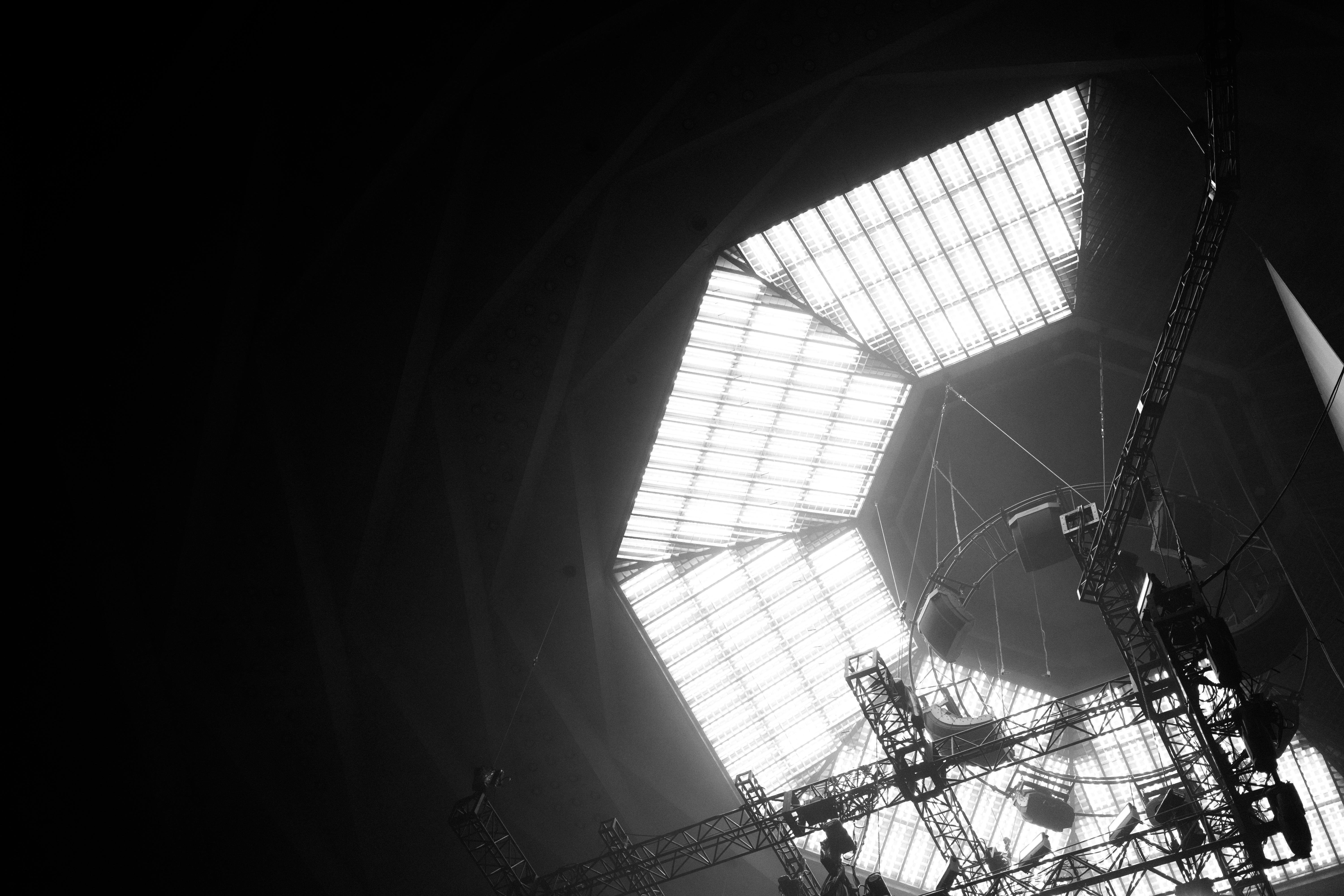 Large skylight ceiling with grid-like panels casting light into a dim interior space. Metal framework with beams and cables visible in the foreground, supporting various equipment. Overall monochrome color scheme with high contrast between light and shadow.