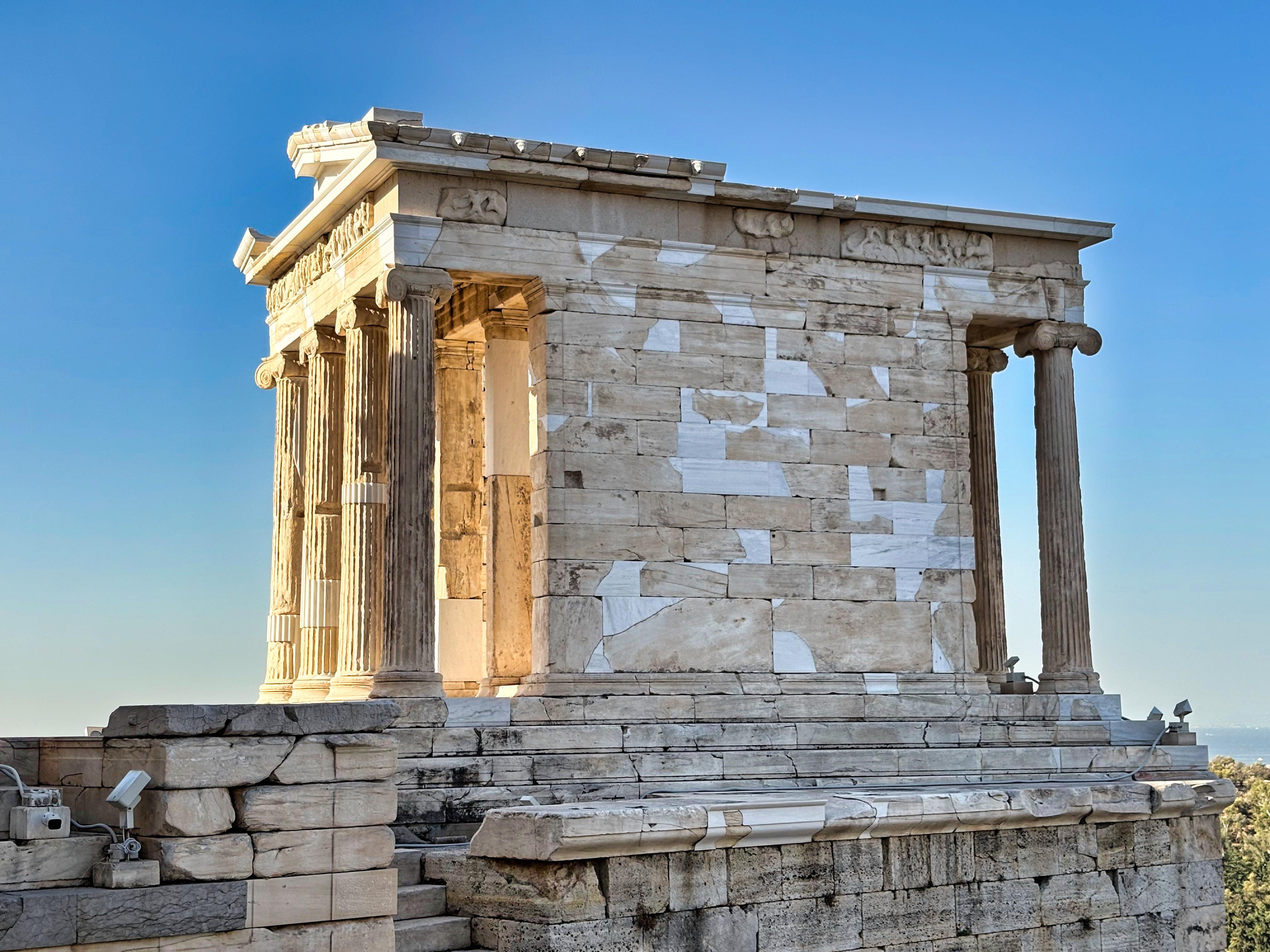 Ancient Greek temple with Doric columns and a pediment, constructed from light-colored stone. The building sits on a stepped platform with clear signs of wear and weathering. The structure has missing sections and irregular patches of restoration. Surrounded by a clear blue sky, indicating a sunny day.
