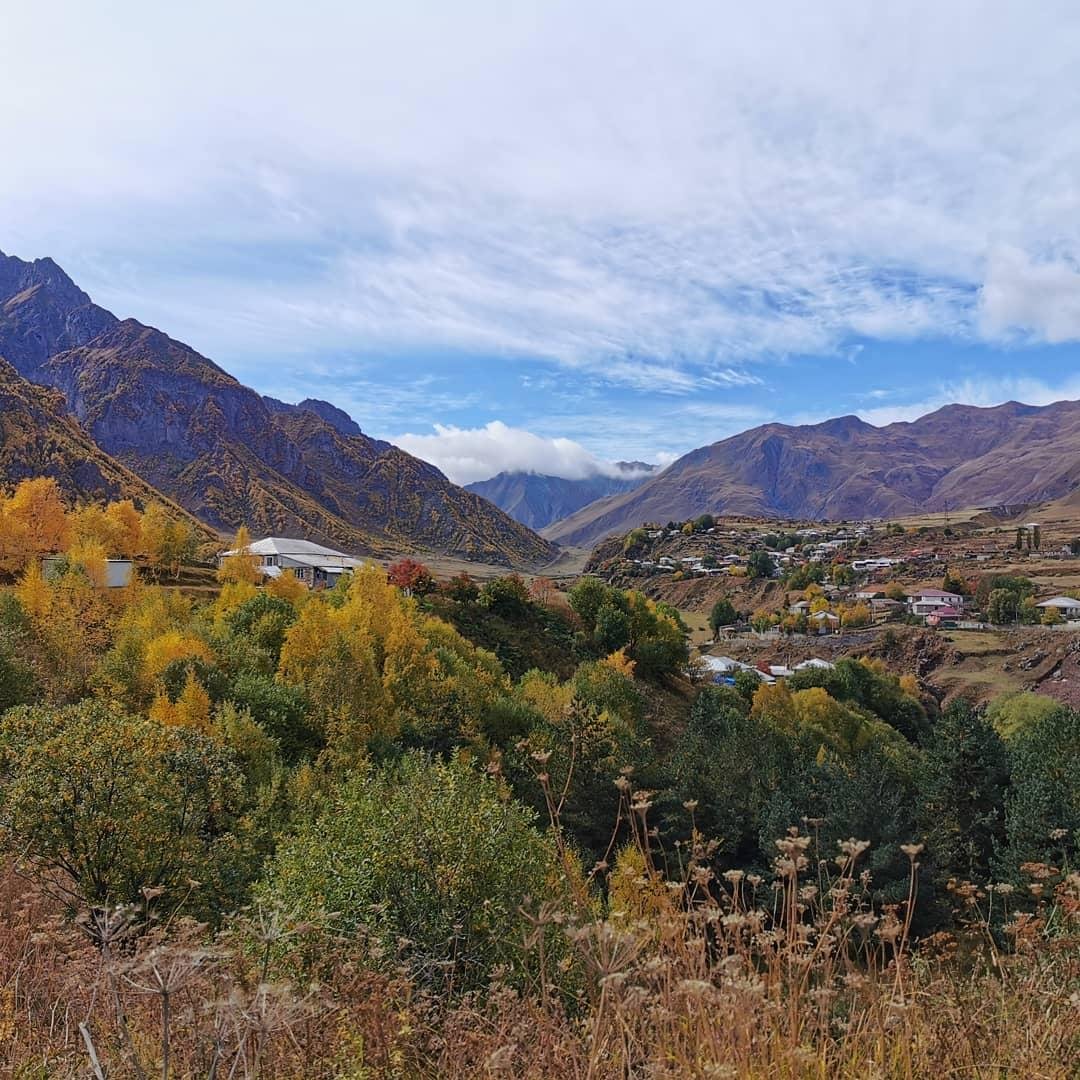Mountain valley landscape with autumn trees in the foreground, a small village of scattered houses on the hillside, rugged peaks in the background, and a partly cloudy blue sky.