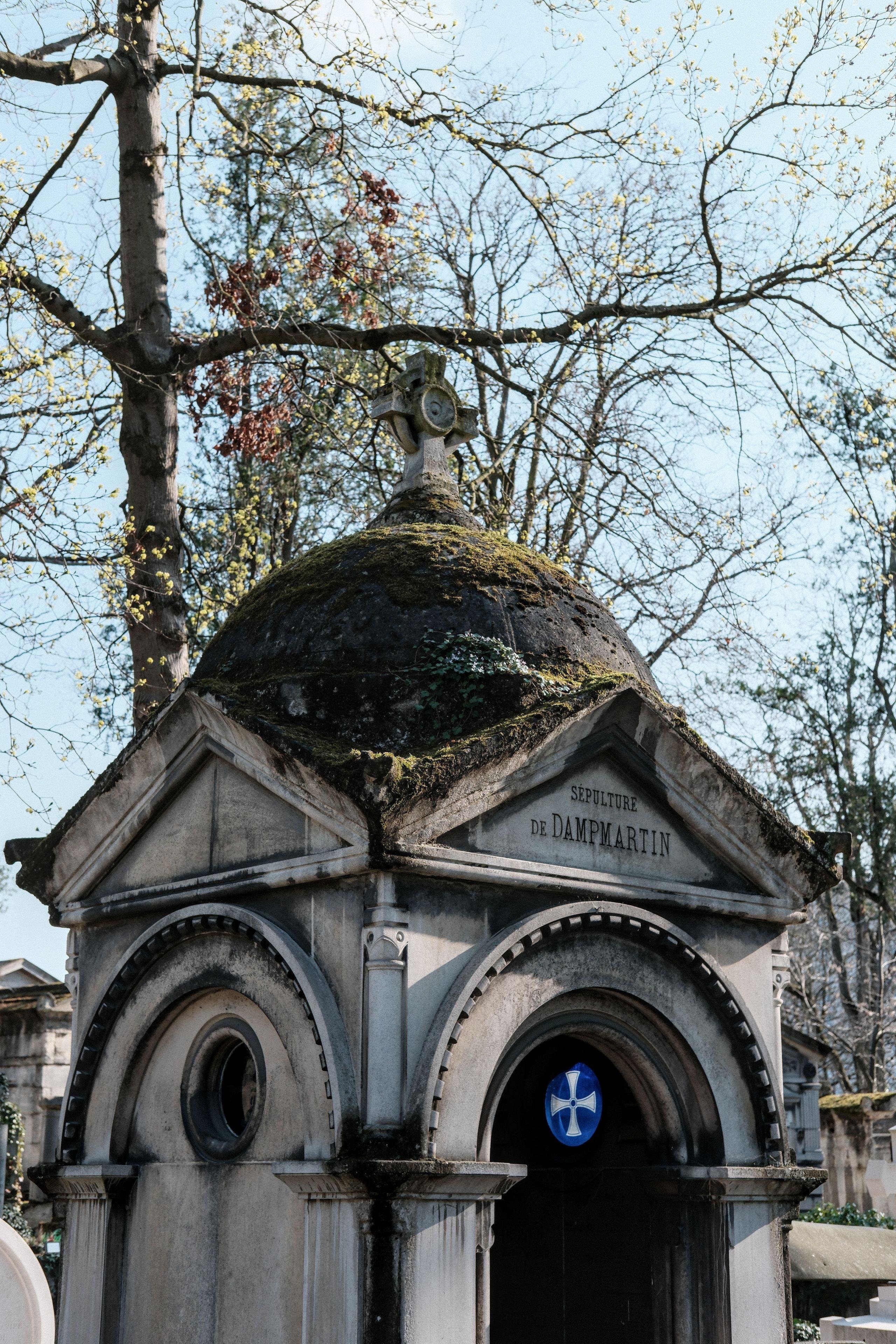 Photo verticale. Une moitié de mausolée à l’intérieur duquel se détache dans le noir un disque bleu lumineux avec une croix régulière blanche dedans. Au dessus du mausolée, des arbres et le ciel bleu