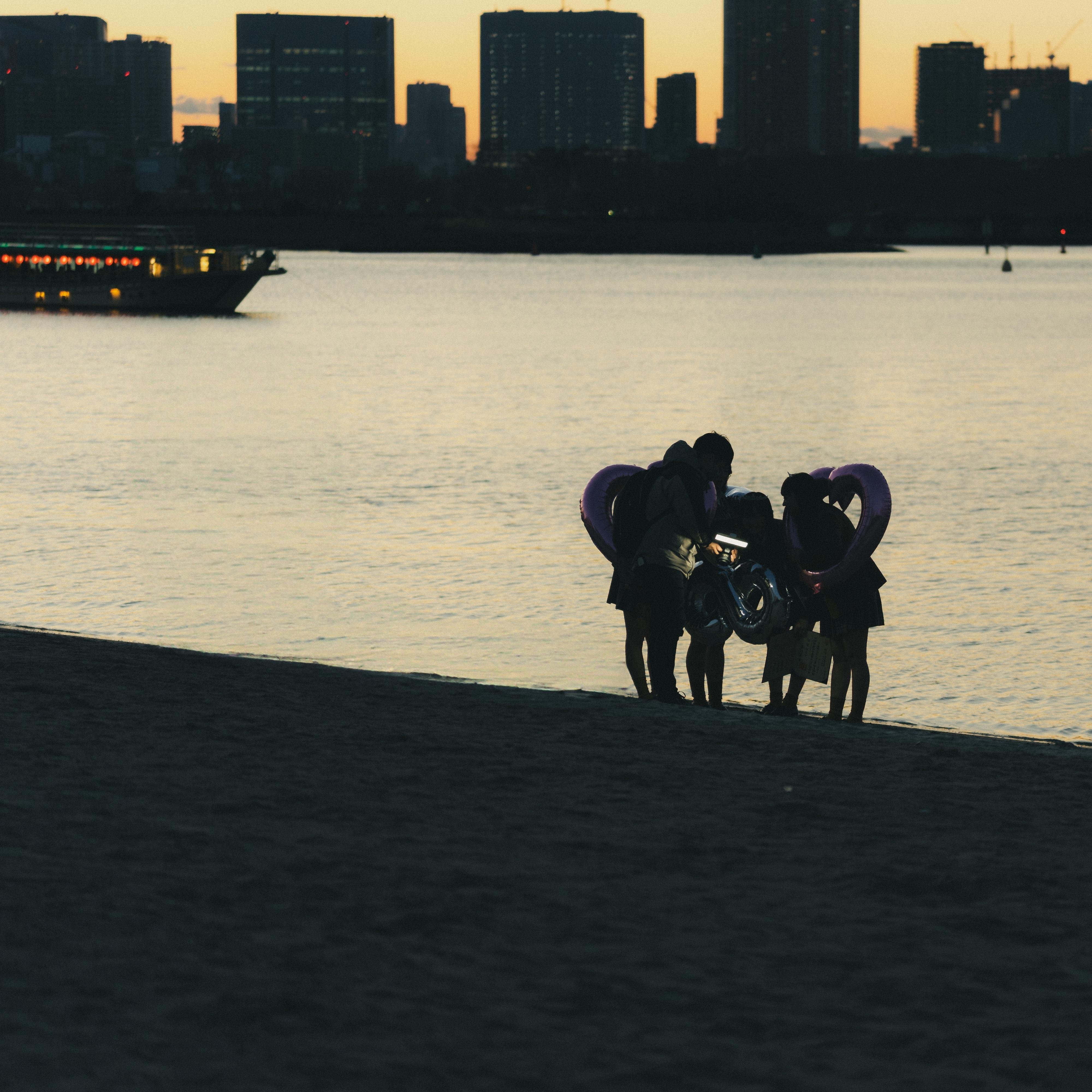 The image depicts a group of people standing near the water's edge at dusk or dawn, with a city skyline in the background. They appear to be holding or surrounded by inflatable rings or balloons, possibly shaped like hearts. The lighting is low, suggesting a sunset or sunrise scene.