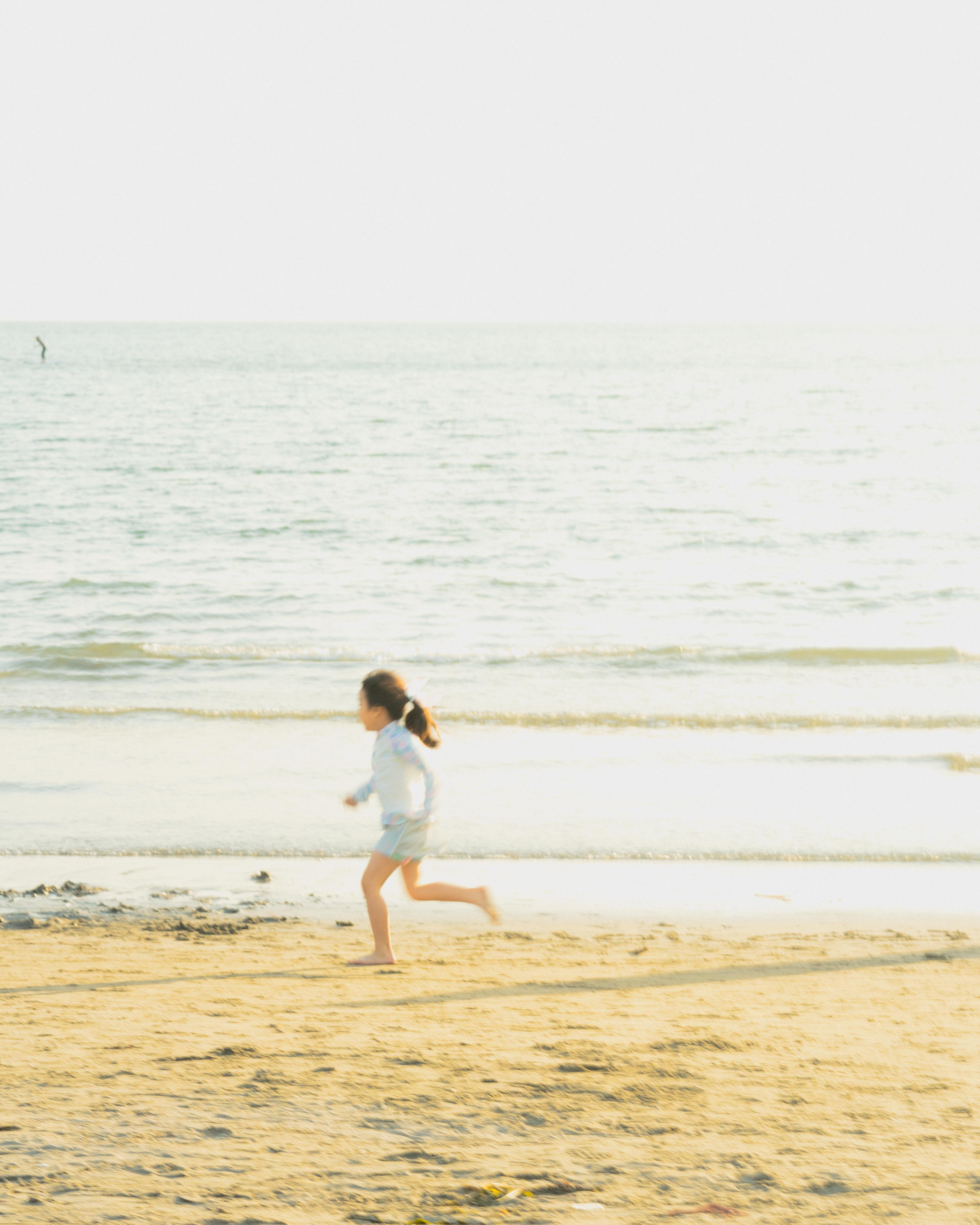 A young girl with a ponytail runs along a sandy beach near the water's edge. The sea is calm with gentle waves and a bright, hazy sky above. A solitary figure is visible in the distant water. The scene is bright and sunny, casting soft shadows on the sand.