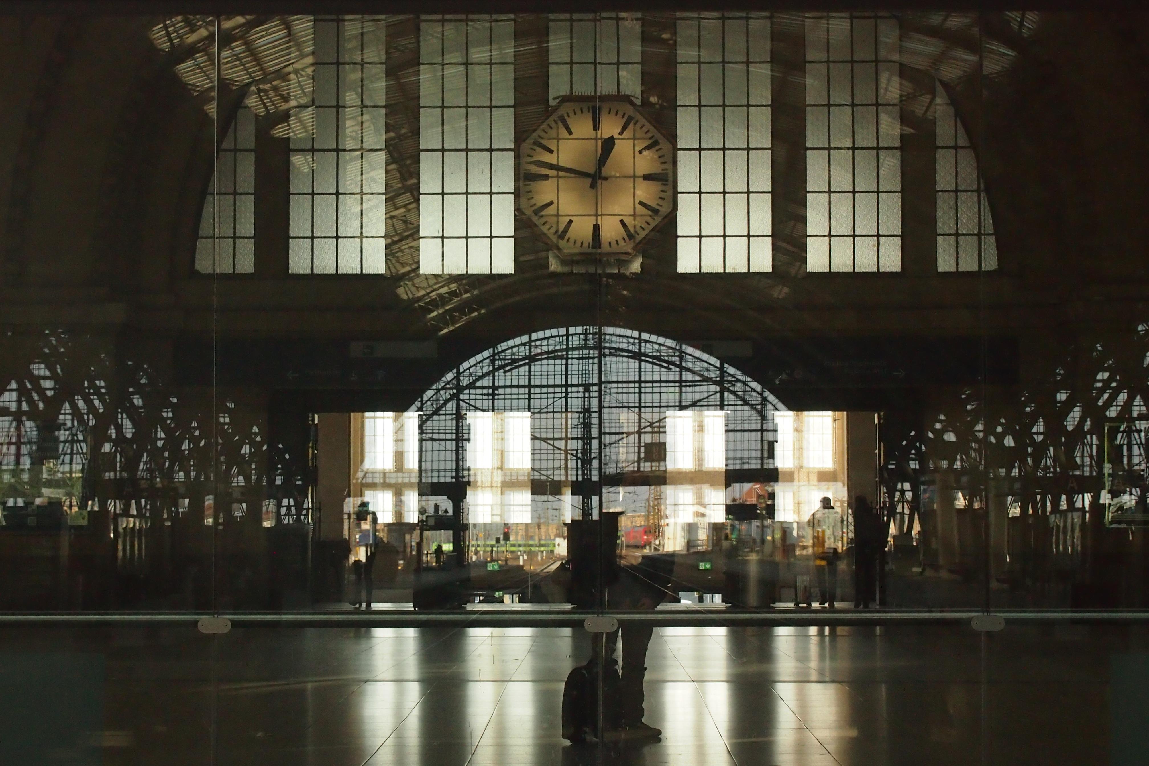 Foto im Querformat.
In einem Bahnhof, Blick auf ein Portal mit großer Uhr oben in der Mitte, daneben Fenster.
Im Vordergrund befindet sich eine Glaswand, in dieser spiegelt sich (von hinter der Kamera) die Gleishalle. Ebenso sind die hintere Wand Richtung Gleisvorfeld mit mehreren Zügen und Tragebögen der Dachkonstruktion zu erkennen.