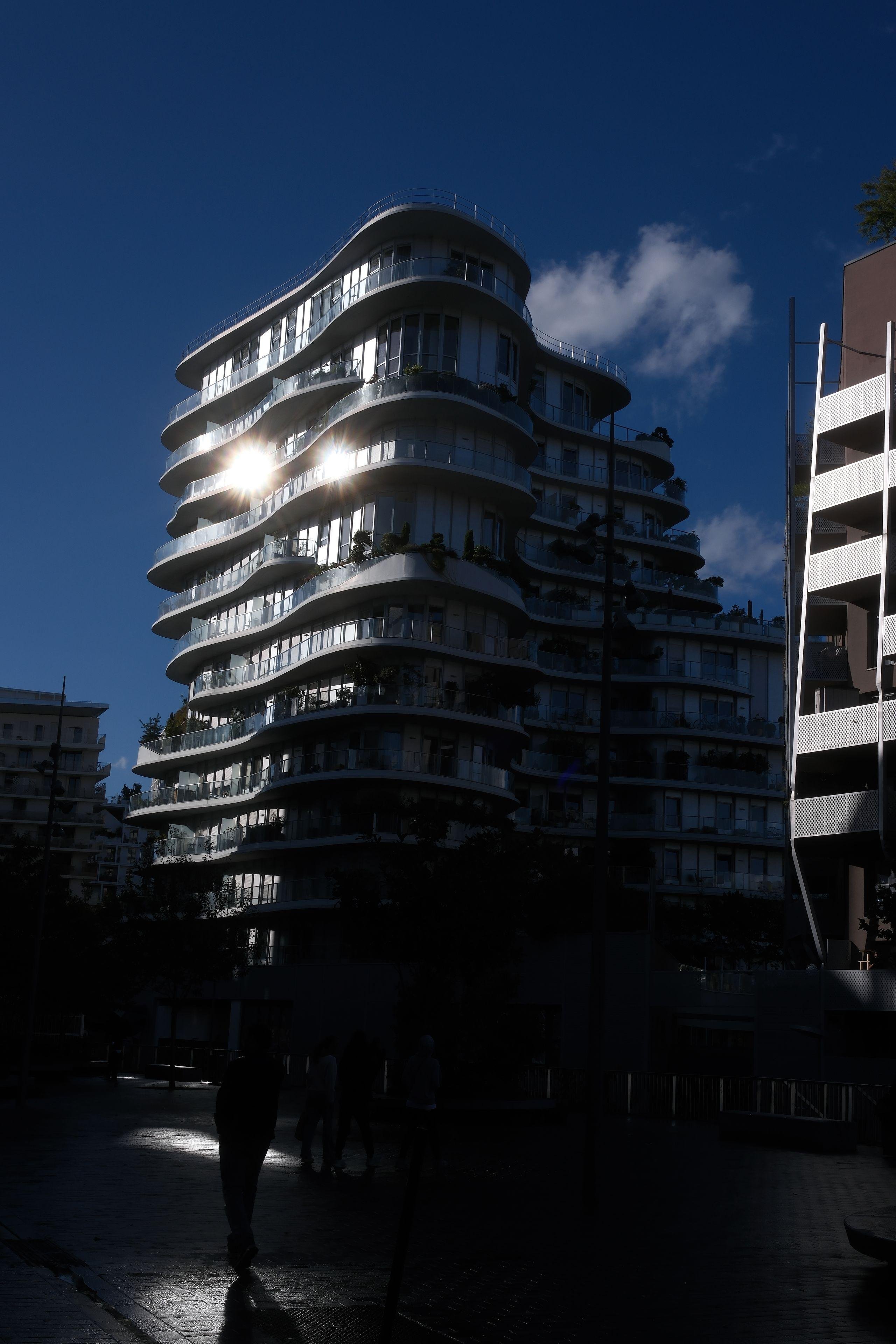 Un immeuble au look très moderne tout en courbes qui réfléchi beaucoup de soleil, rendant le premier plan tout sombre, dans laquelle une silhouette se devine, le tout sous un ciel bleu