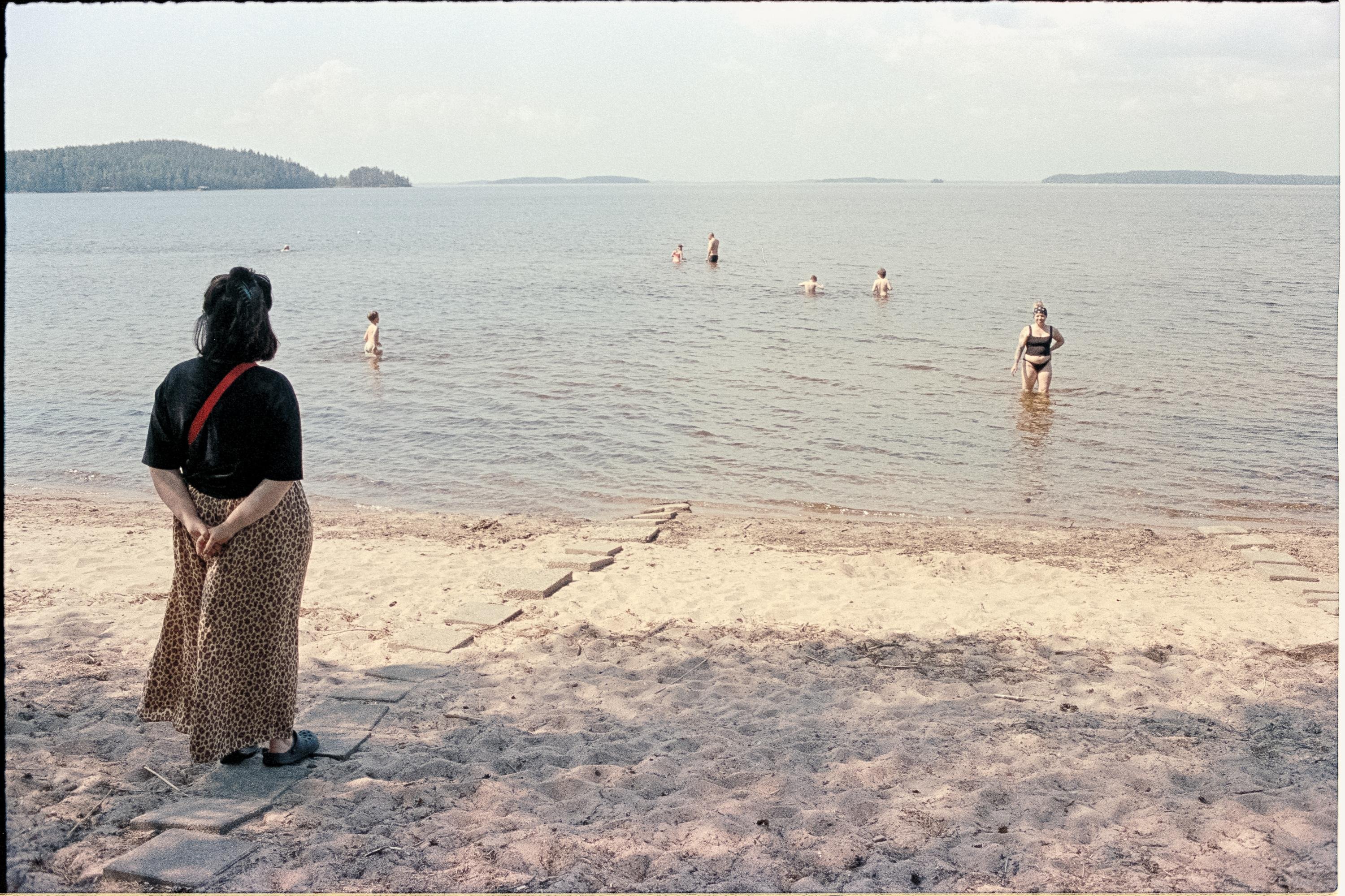 A person in a black shirt and leopard-print skirt stands at the edge of a sandy beach, facing a calm body of water. Several people are swimming in the water, some closer to the shore and others farther out. The background features distant forested land across the water under a partly cloudy sky.