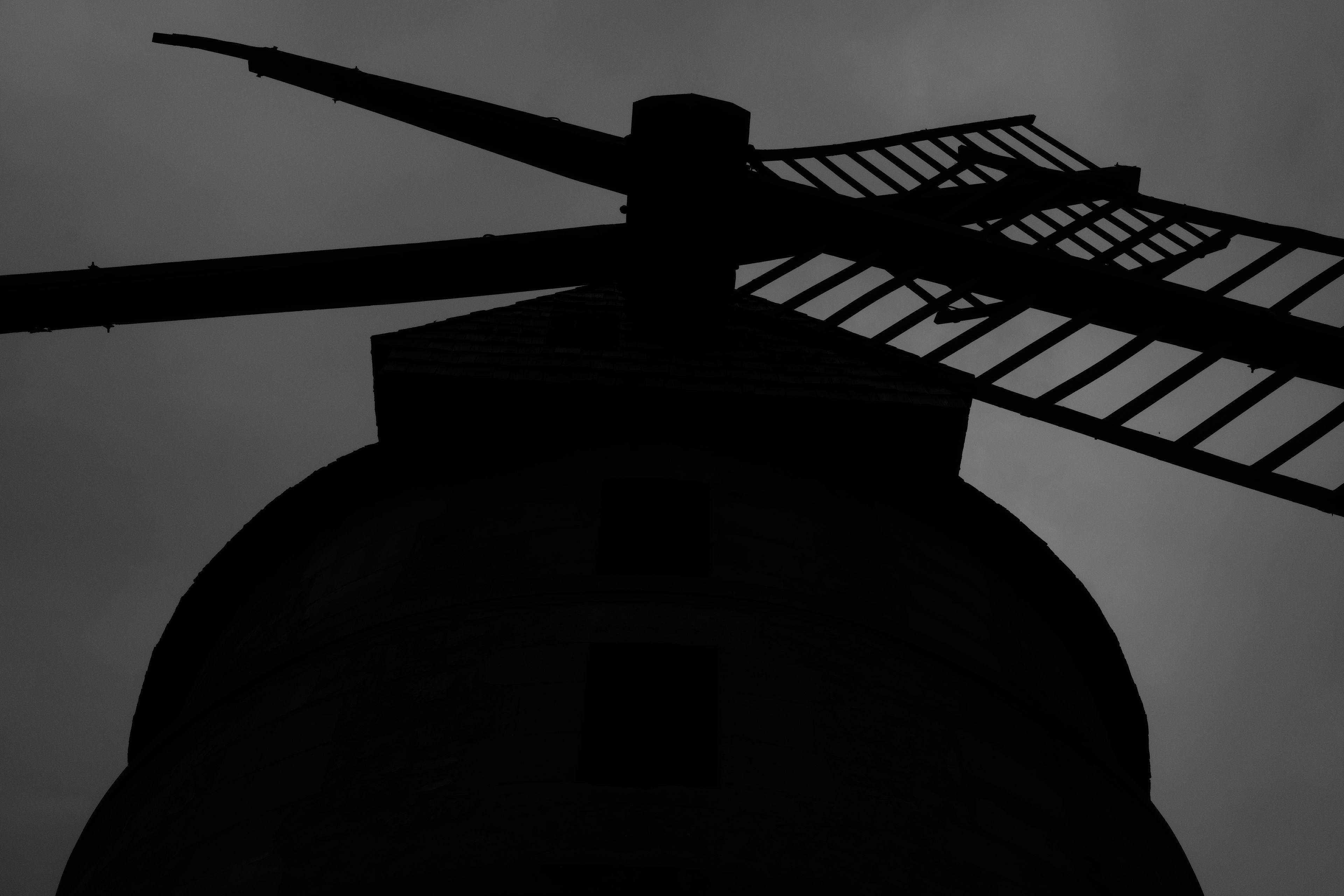 Photo noir et blanc en contre plongée d’un moulin à vent, de face, en contre jour, sur un ciel gris, donnant une impression menaçante à l’ensemble