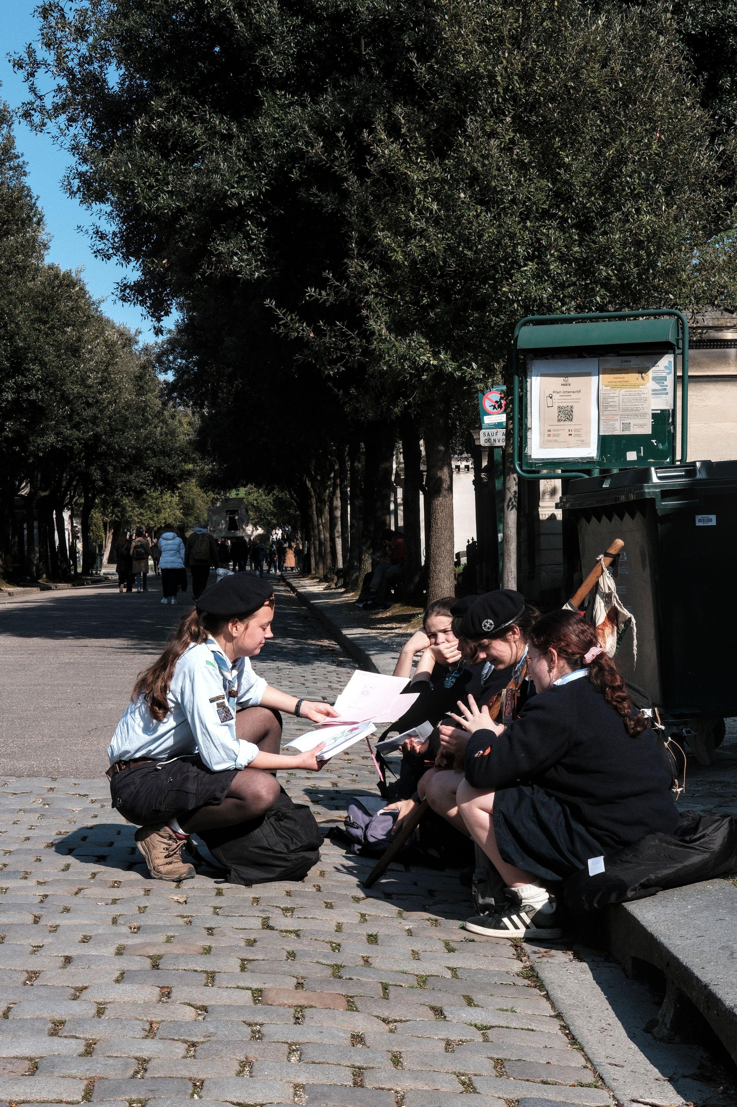 Photo verticale. Un groupe de filles scouts assises sur le bord d’un trottoir, une autre portant un berêt accroupie devant elles leur tendant des feuilles.
