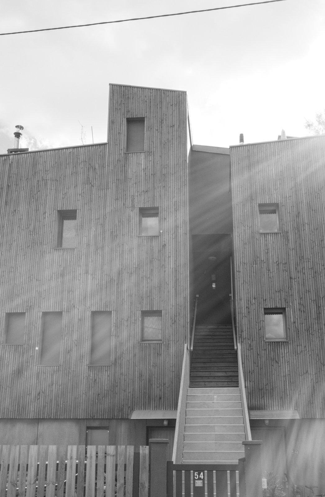 Photo en noir et blanc de la façade d’un immeuble de forme géométrique sortant de l’ordinaire, asymétrique, avec une façade en bois. Un flare de lumière éclaire la photo par l’angle supérieur droit